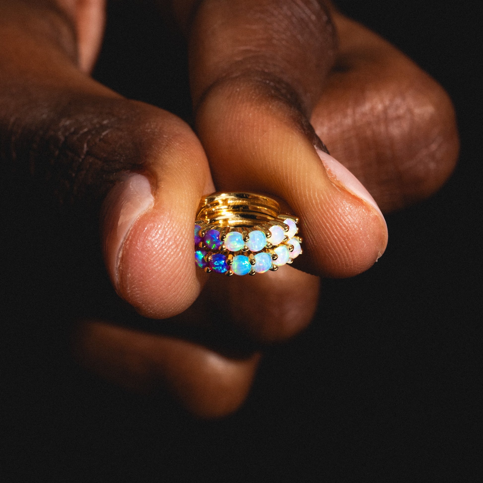 A hand displays the Mini Opal Ombre Huggies—gold huggie earrings set with multi-colored opals, sparkling against a dark background, and crafted in radiant 18k yellow gold vermeil.