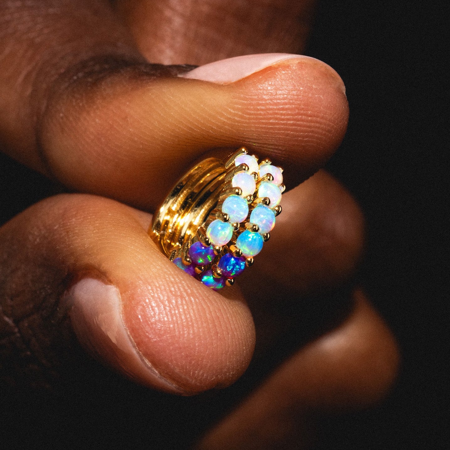 A close-up of a hand holding the Mini Opal Ombre Huggies, crafted in 18k yellow gold vermeil and set with lab-created opals that catch the light against a dark backdrop.