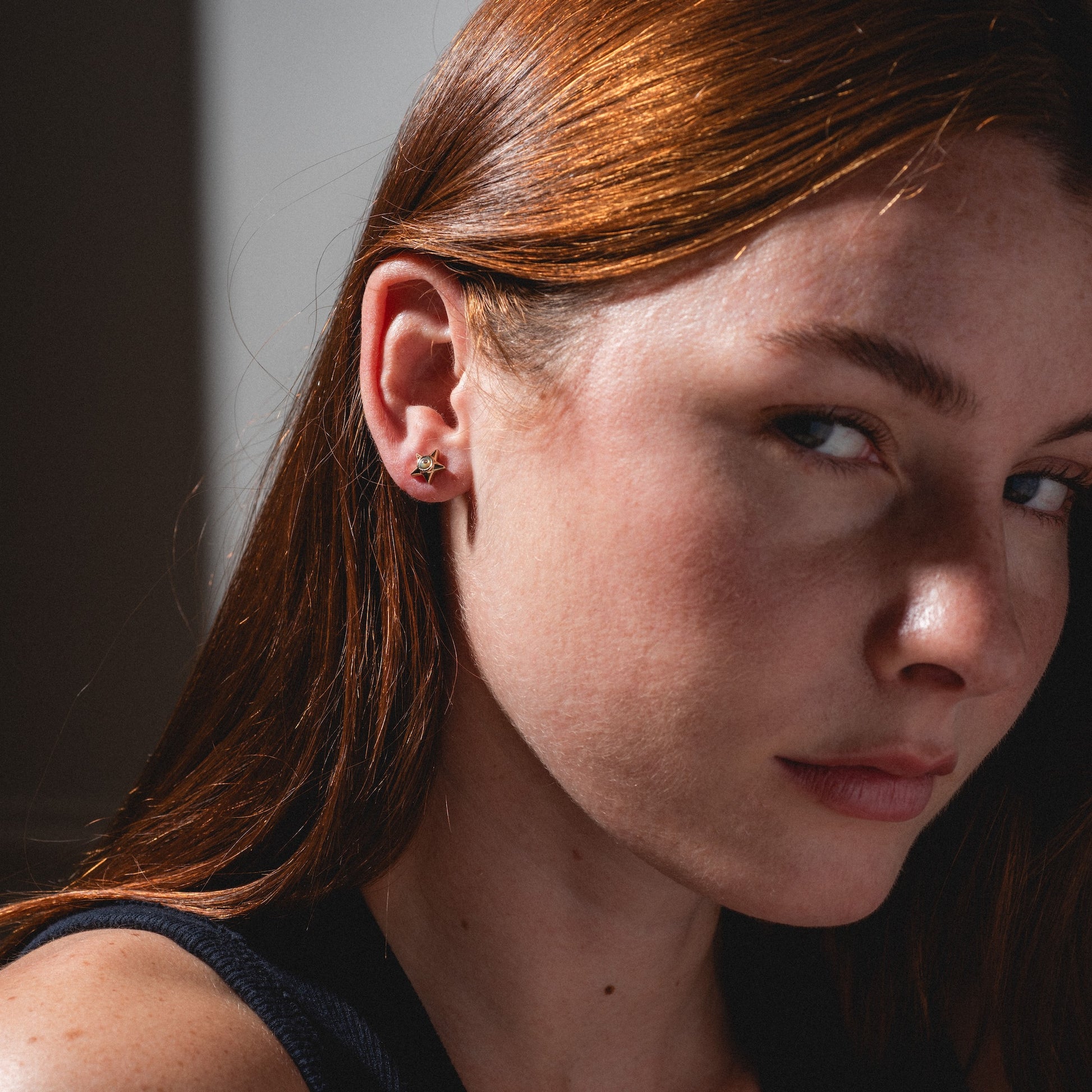 Close-up of a woman with long red hair wearing the Lucky Star and Moon Studs, looking slightly past the camera with a neutral expression. Sunlight highlights her face and hair.