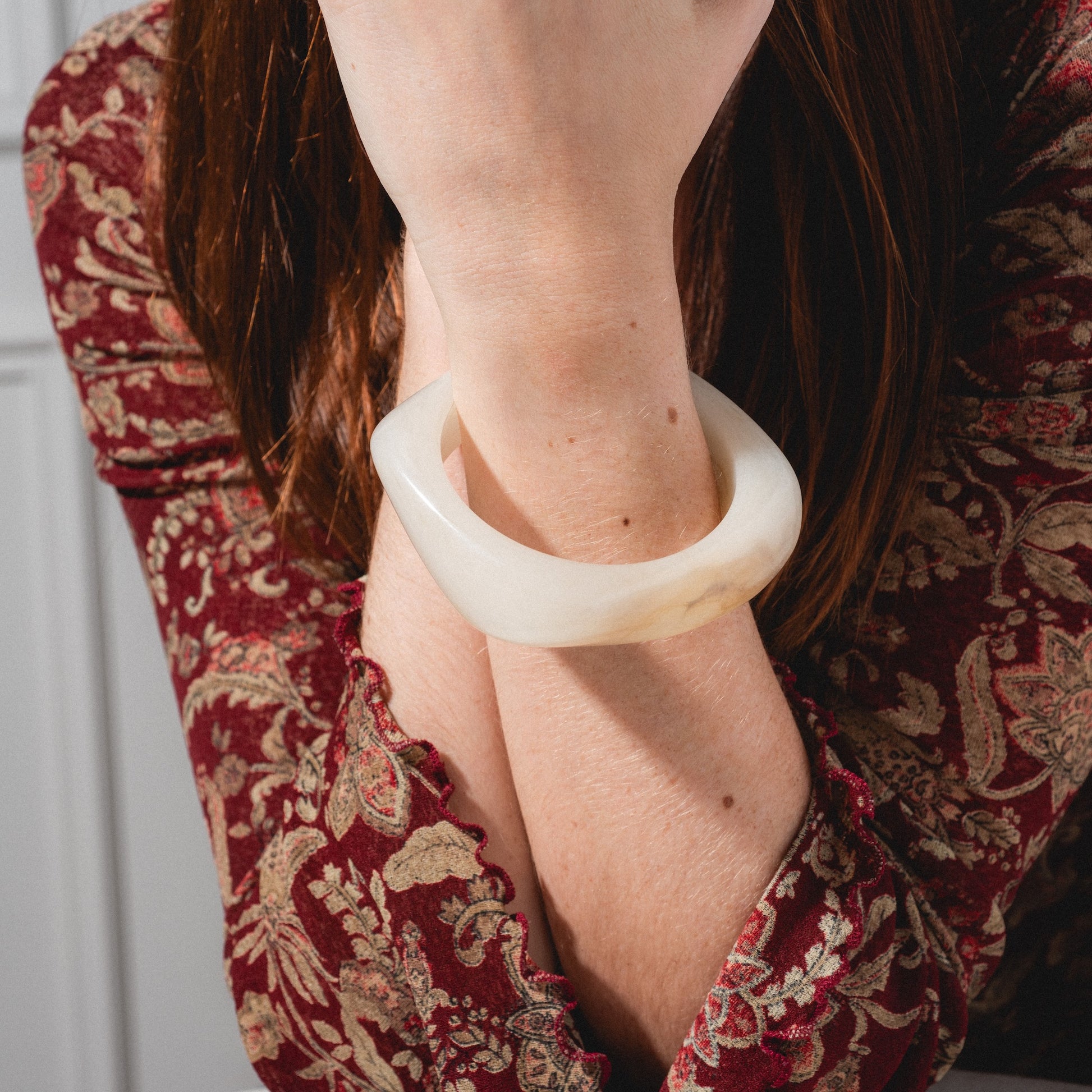 A person in a paisley burgundy top rests their chin on their hand, displaying the Bangle Bracelet on their wrist. Their face is partly hidden by their hand and hair.