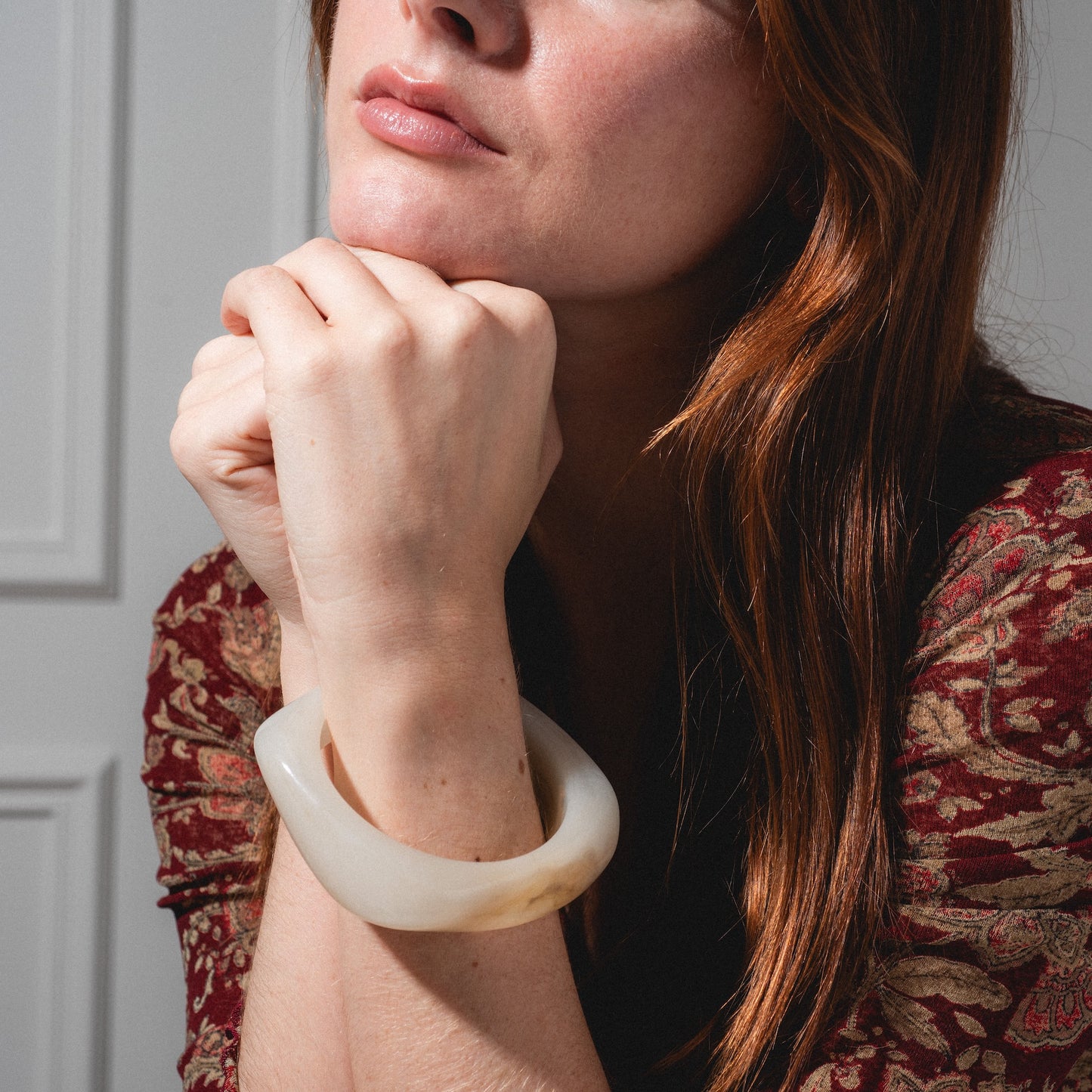 A woman with long red hair, dressed in a maroon and gold patterned top, rests her chin on her hands while wearing the Bangle Bracelet. She is posed in soft natural light.