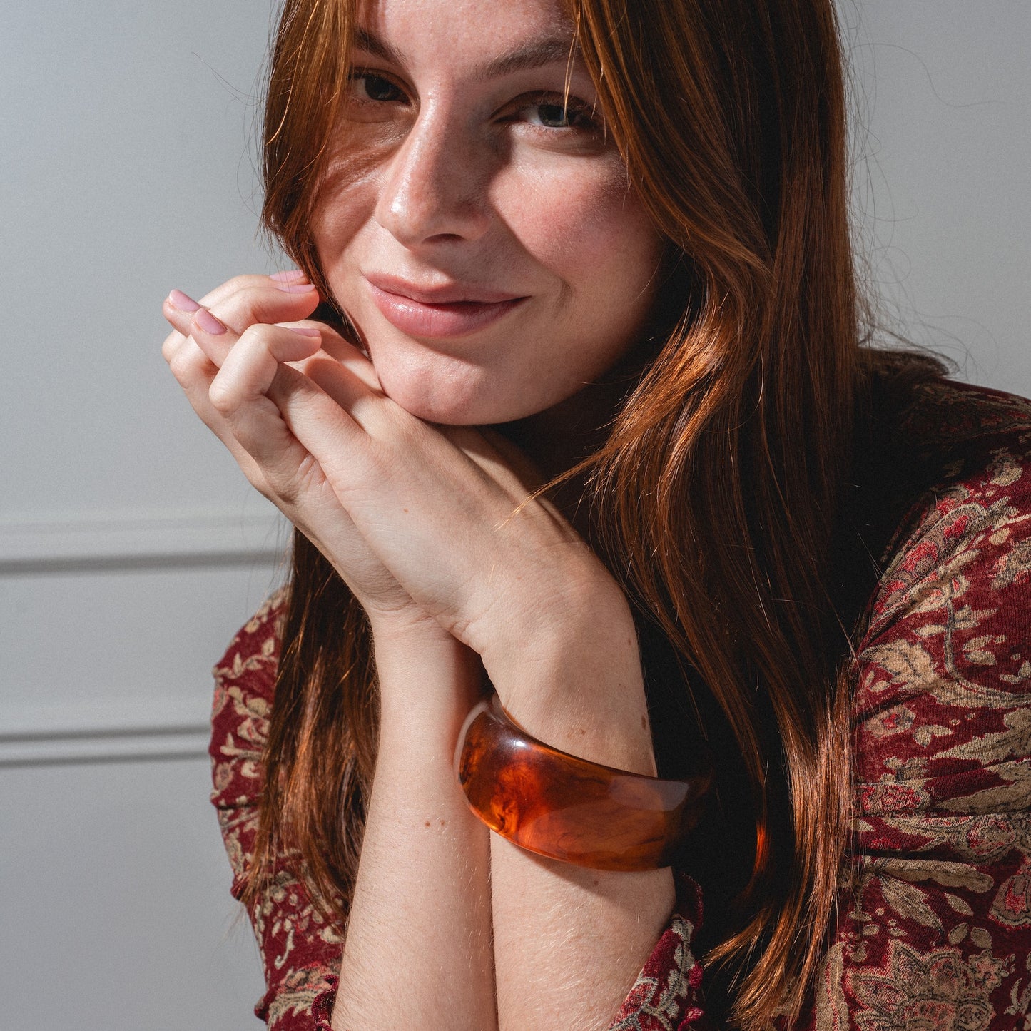 A woman with long auburn hair and fair skin smiles gently, resting her chin on her hands. She wears a patterned red top and completes her look with the Bangle Bracelet. The background is light and minimal.
