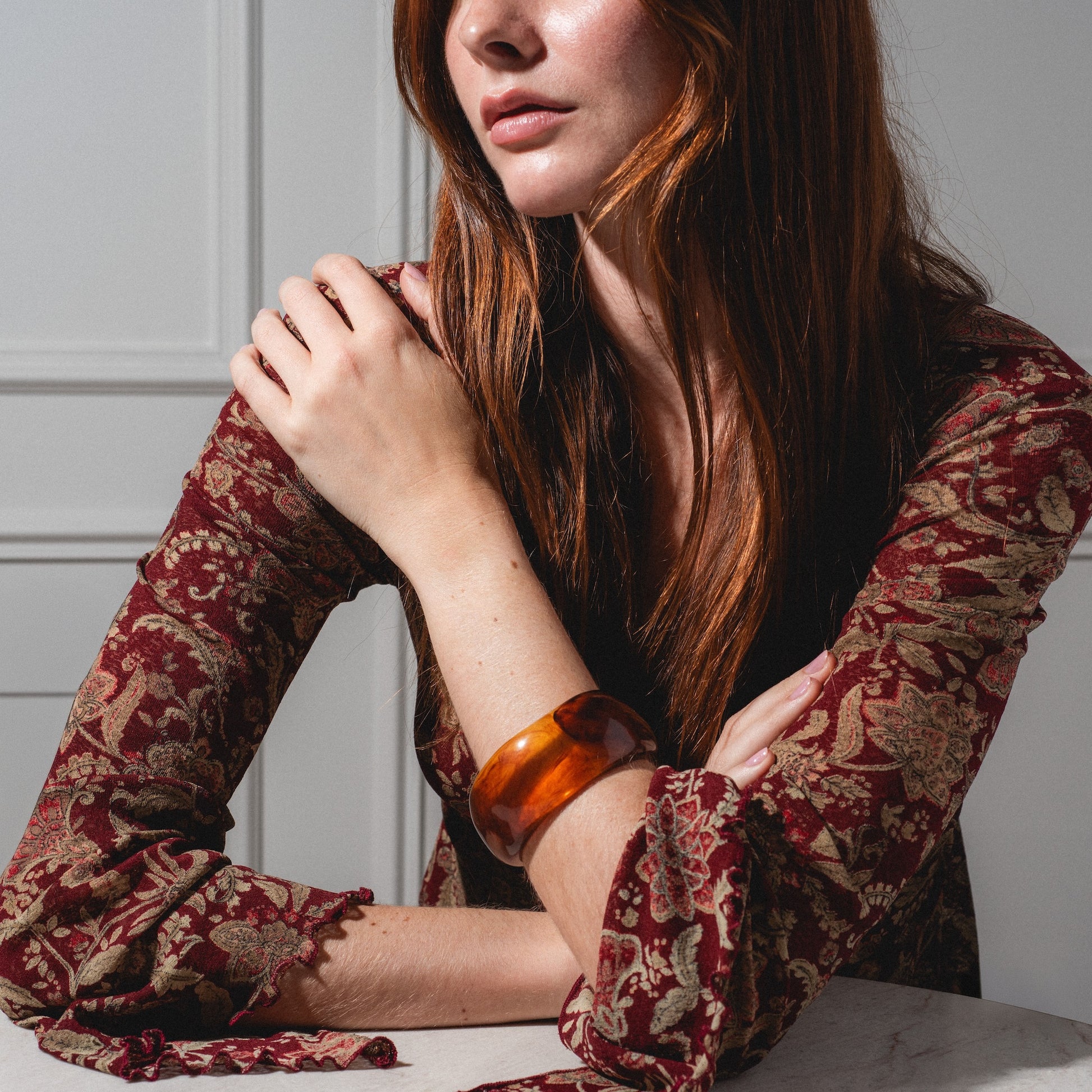 A woman with long red hair, wearing a patterned burgundy and gold top, sits at a table. She has a large Bangle Bracelet on her wrist and rests one arm across her chest. Her face is partially visible.