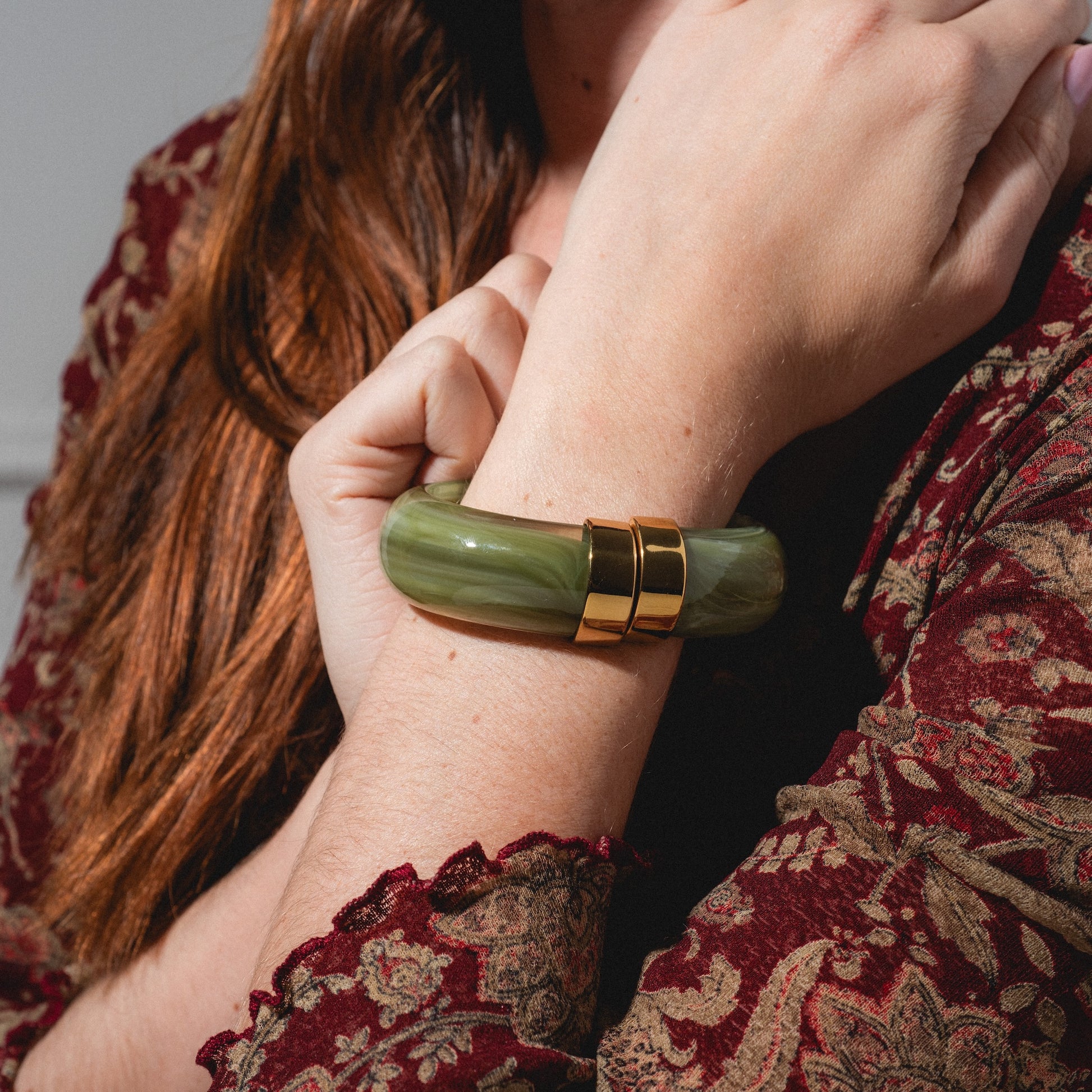 A person in a patterned burgundy sleeve displays their wrist adorned with the Sylune Bangle Bracelet, a chunky green hinged piece accented with two gold metal details, as their hand rests gently on their opposite shoulder.