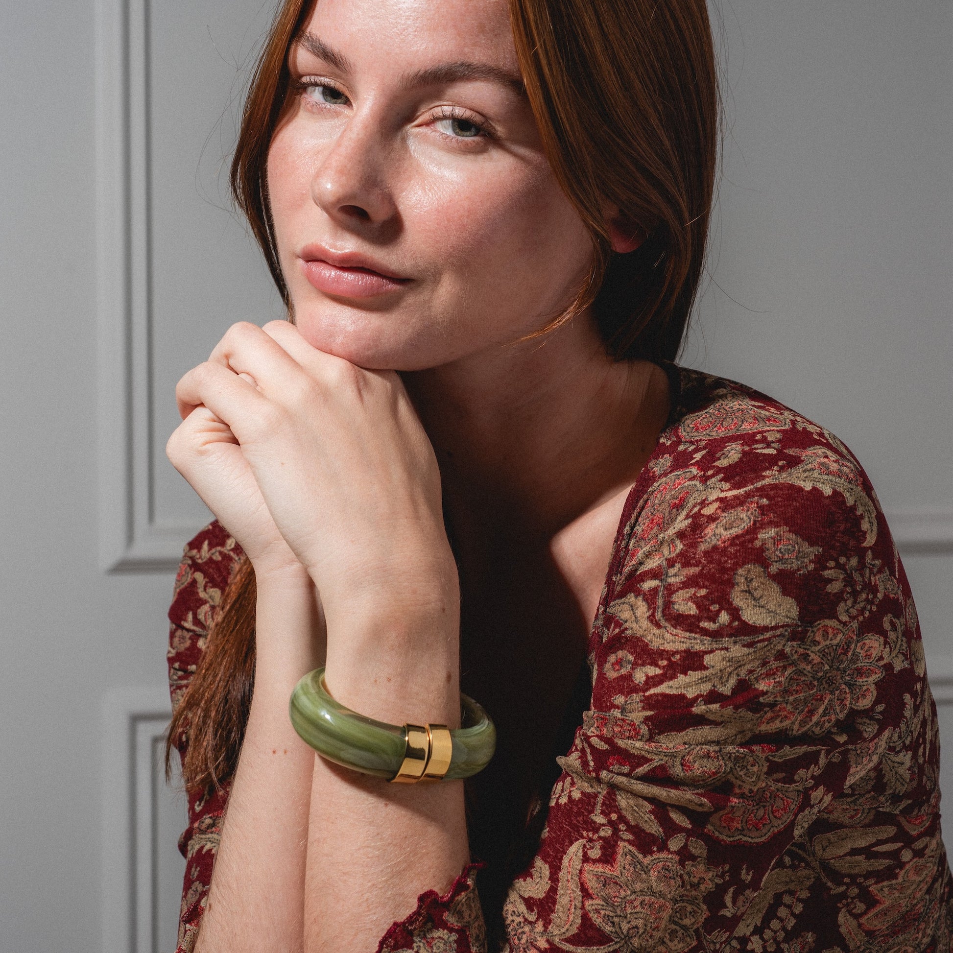 A woman with long red hair, in a red patterned top and wearing the Sylune Bangle Bracelet, rests her chin on her hands and gazes softly at the camera against a neutral background.