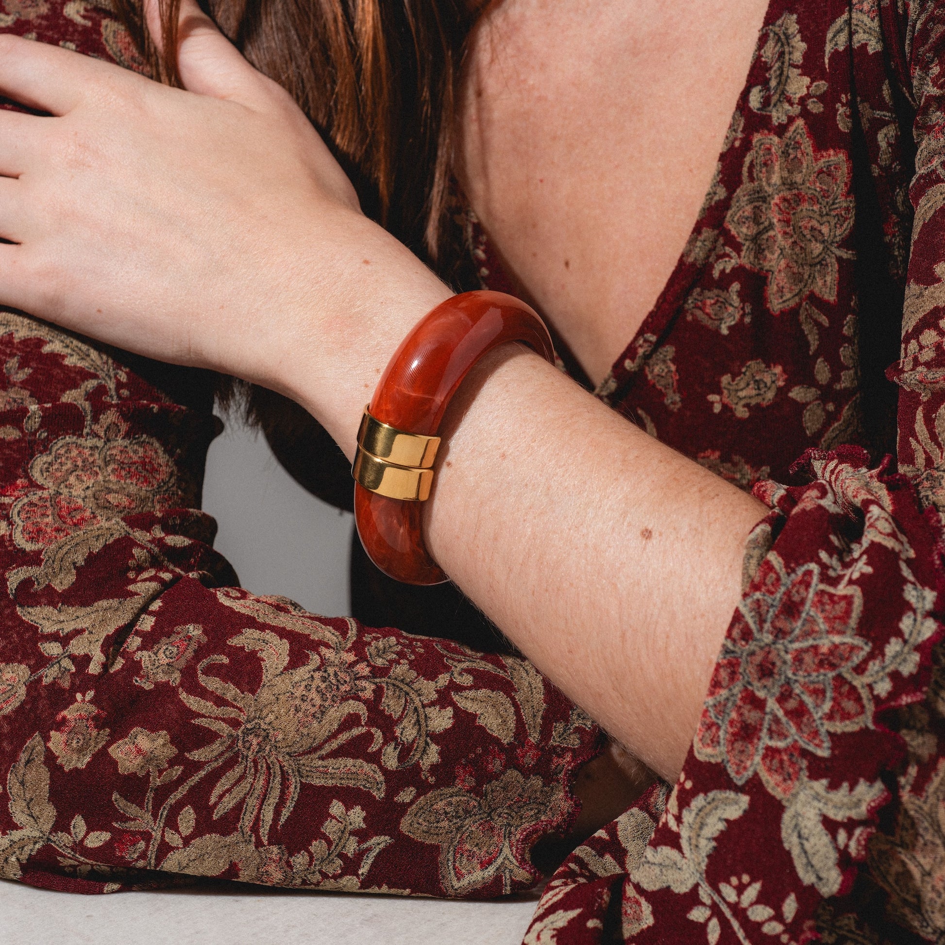 A person wearing a maroon floral long-sleeve top rests their arm on a surface, showcasing the Sylune Bangle Bracelet—a chunky red and gold hinged accessory—on their wrist.