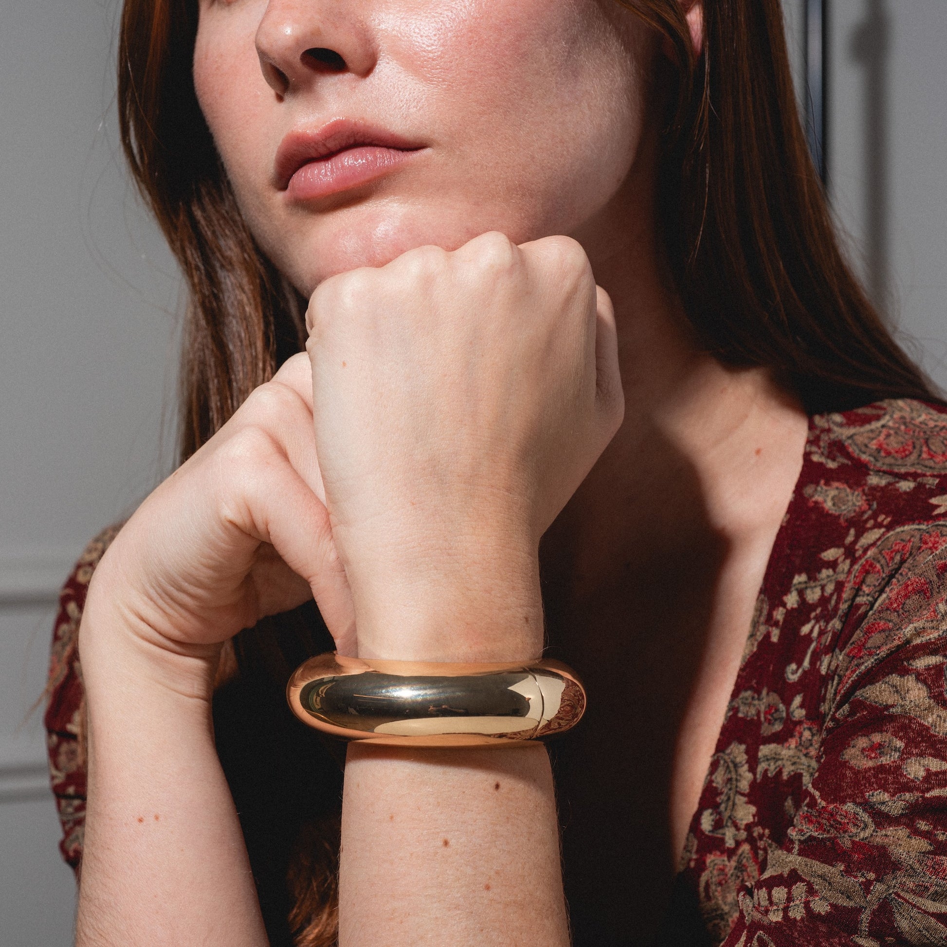 A woman with long reddish-brown hair rests her chin on her fist, displaying the shiny, thick Bangle Bracelet on her wrist. She is dressed in a short-sleeved, patterned burgundy top.