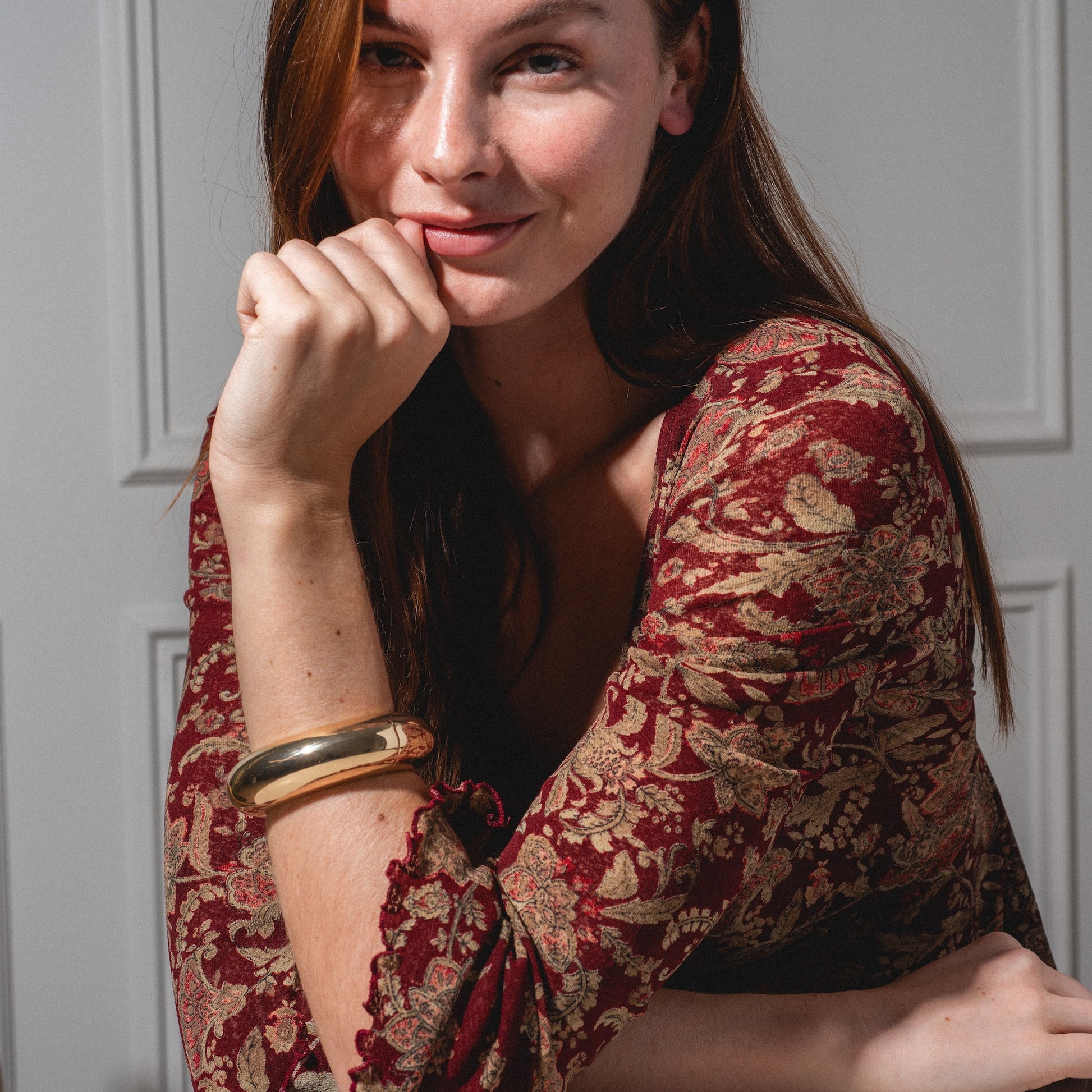 A woman with long brown hair smiles softly indoors, wearing a patterned red and beige top and the Bangle Bracelet on her wrist, her hand resting on her chin against a white paneled wall.