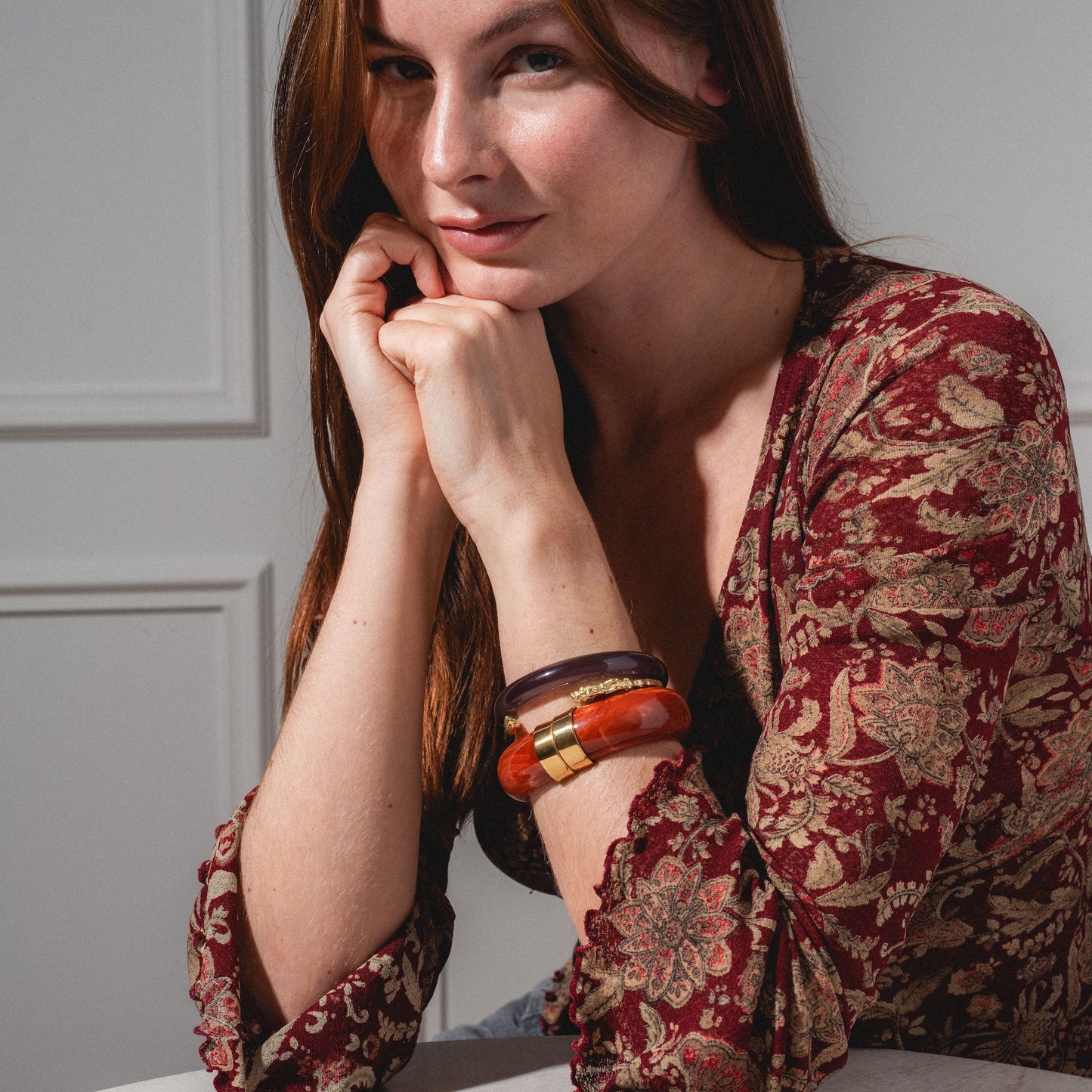 A woman with long brown hair wears a patterned red and beige top and chunky bracelets, including the 22.4 mm Sylune Bangle Bracelet, resting her chin on her hands and smiling softly in natural light.