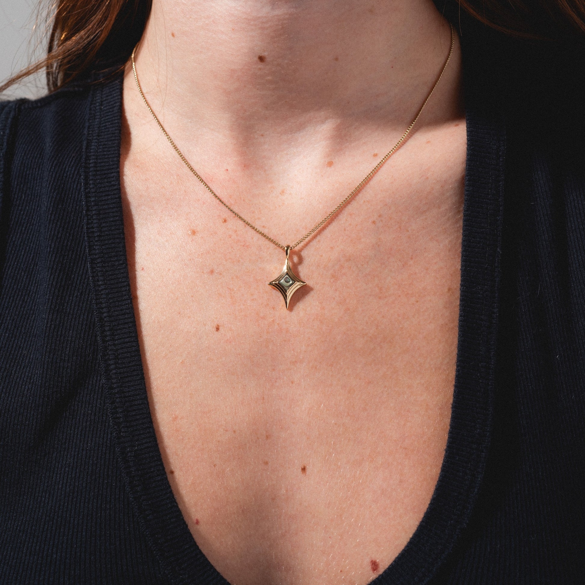 A close-up of a person wearing a deep v-neck black top and the Stellina Pendant Necklace, featuring a gold chain with a diamond-shaped pendant and small central stone, with focus on the necklace and neckline.
