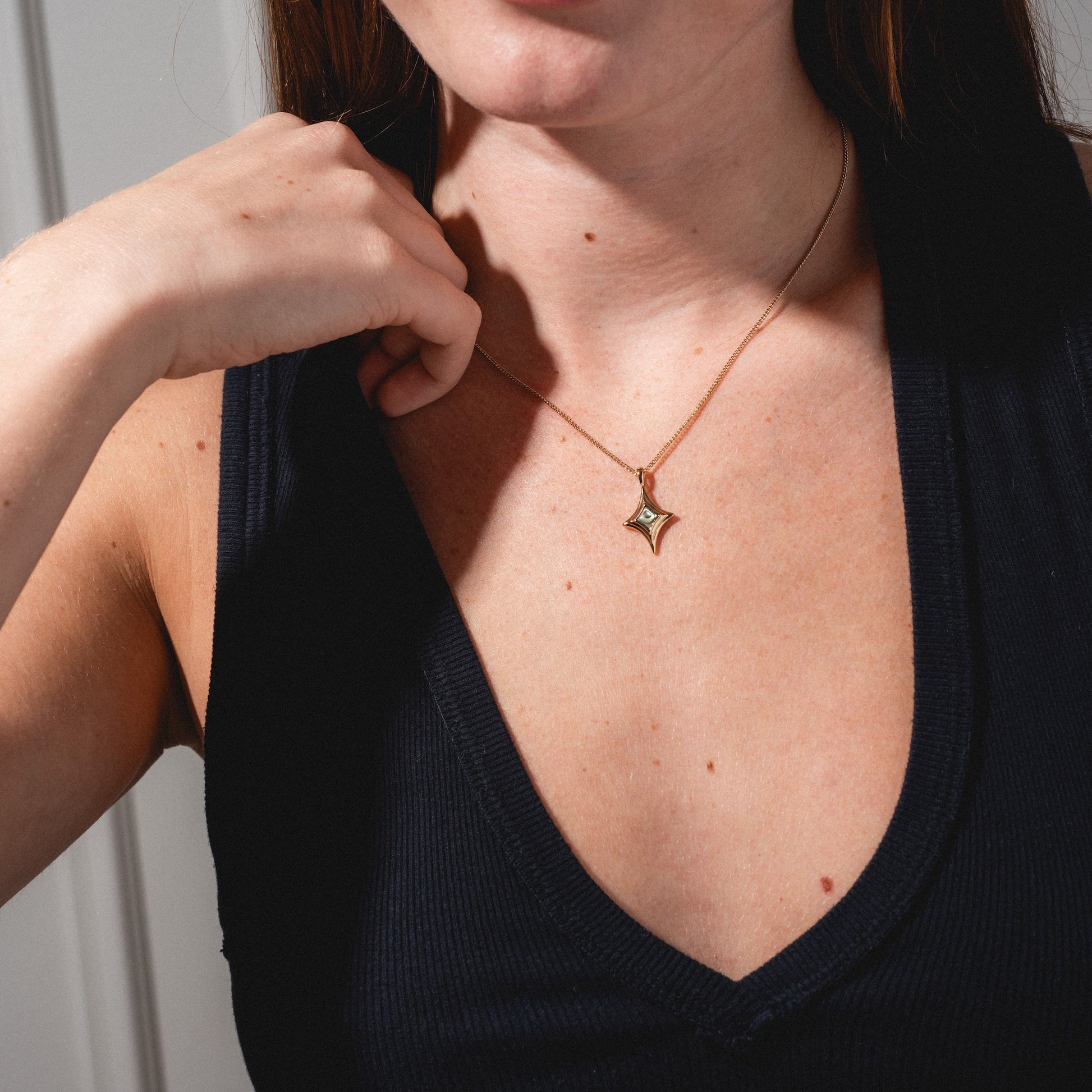 A woman in a sleeveless black top displays the Stellina Pendant Necklace, featuring a star-shaped charm. The image highlights her chest and hand, with natural light emphasizing the elegance of the jewelry.
