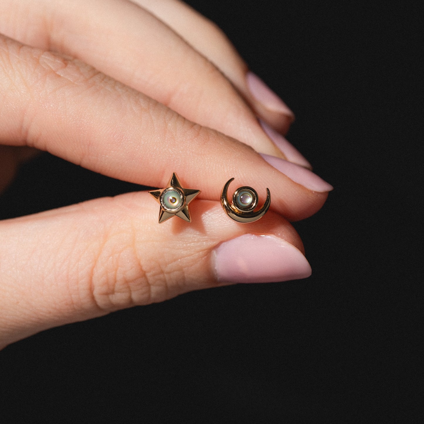 A hand with light pink nail polish holds the Lucky Star and Moon Studs—gold earrings shaped as a star and a crescent moon, each adorned with a tiny central pearl—against a black background.
