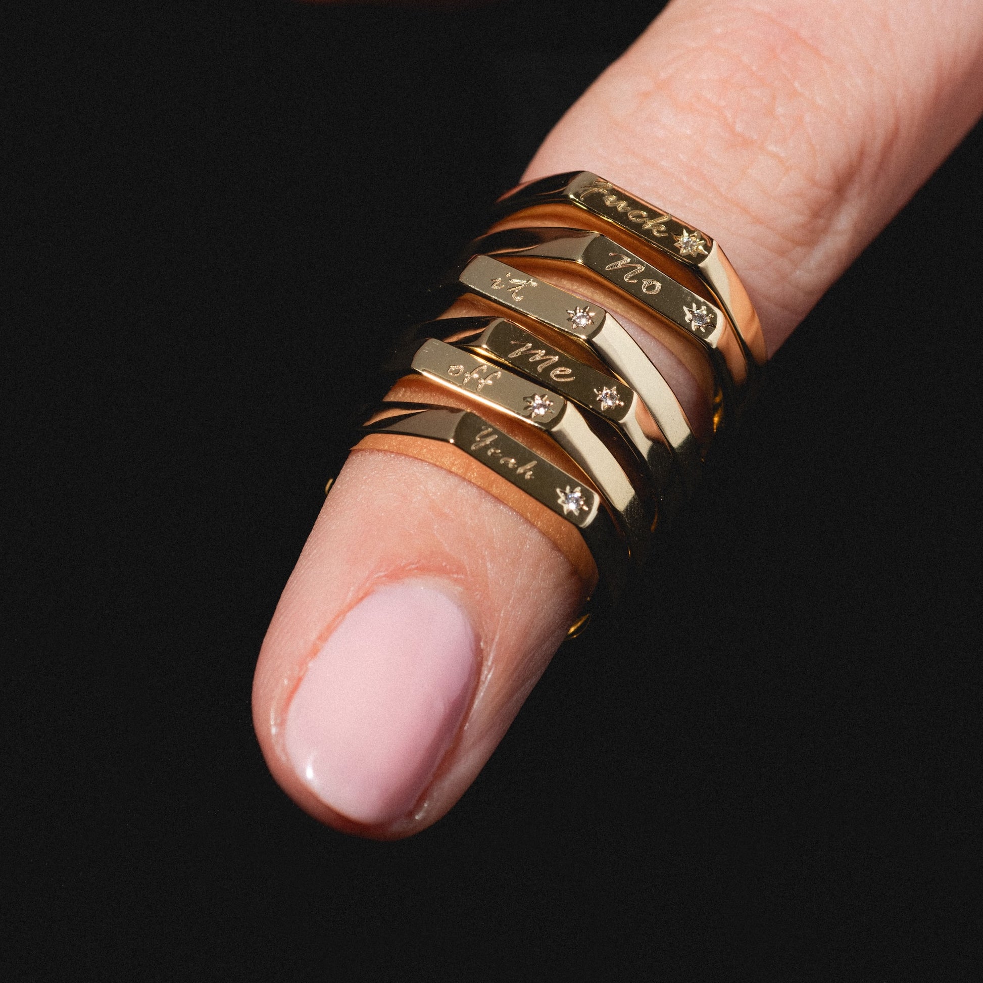 A close-up of a finger wearing the Tiny Conversation Stacking Ring—six gold rings engraved with small text and tiny gems—paired with light pink nail polish, set against a black background.