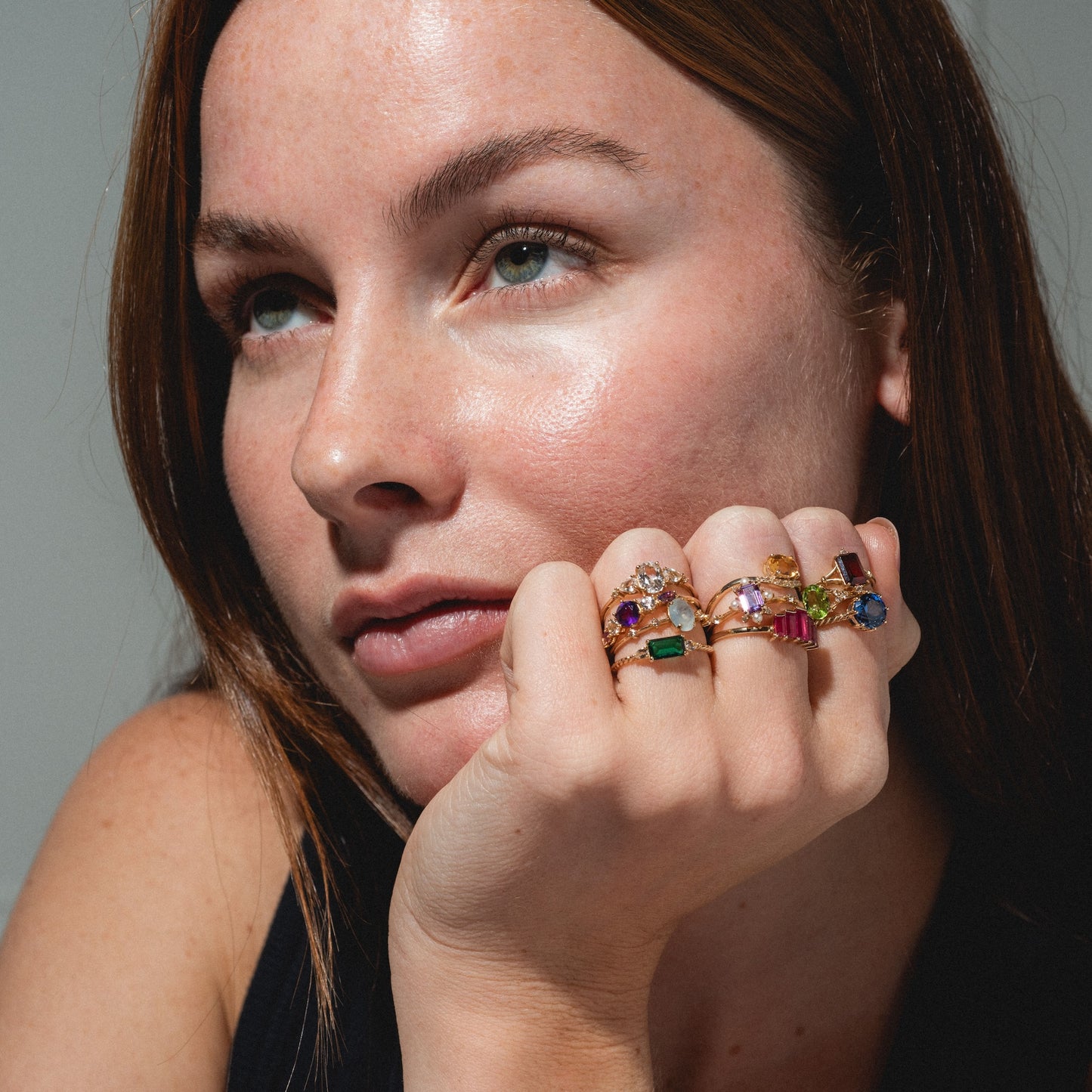 A fair-skinned woman with brown hair gazes thoughtfully to the side, resting her chin on her hand and wearing the Solid Gold 2025 February Capsule Garnet Full Set rings, each featuring colorful gemstones.