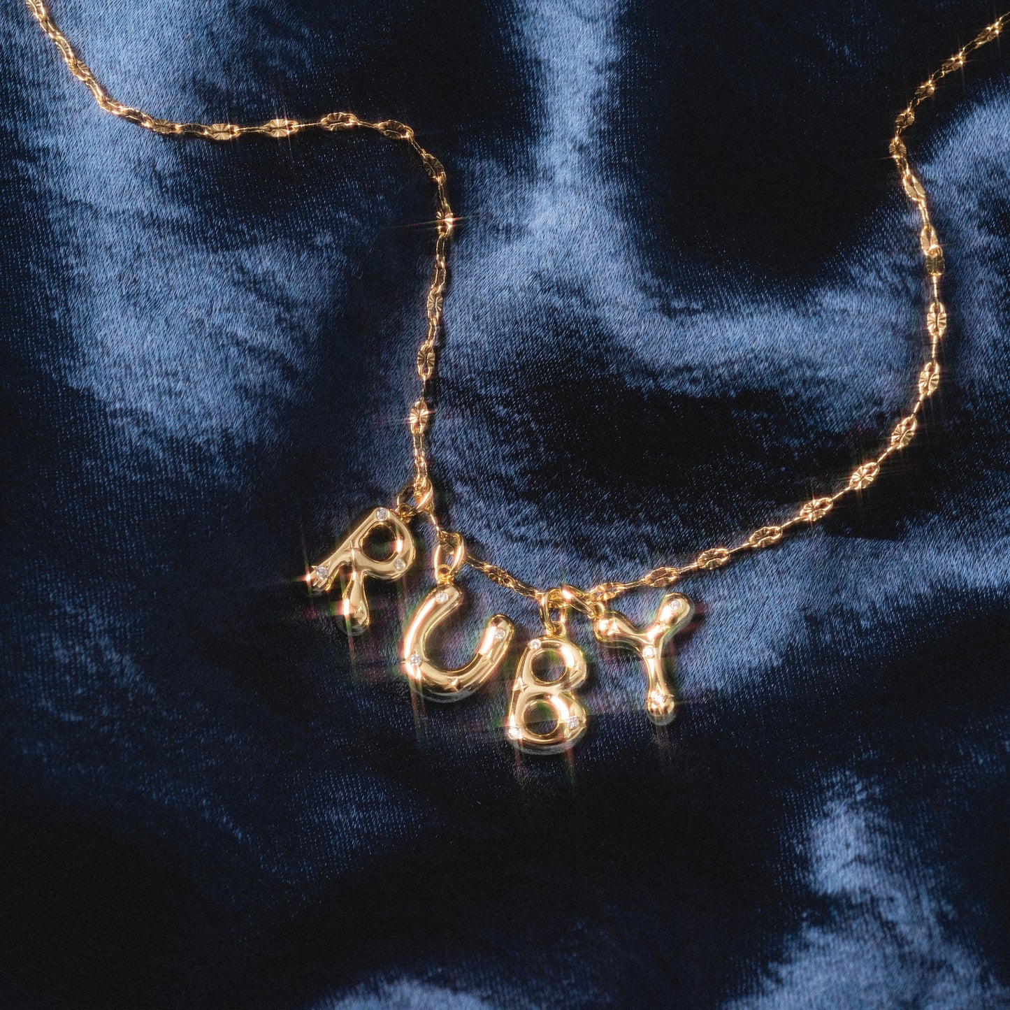 A Star Crossed Celestial Bubble Letter Charm spelling RUBY rests on a dark blue, shiny, textured fabric background.