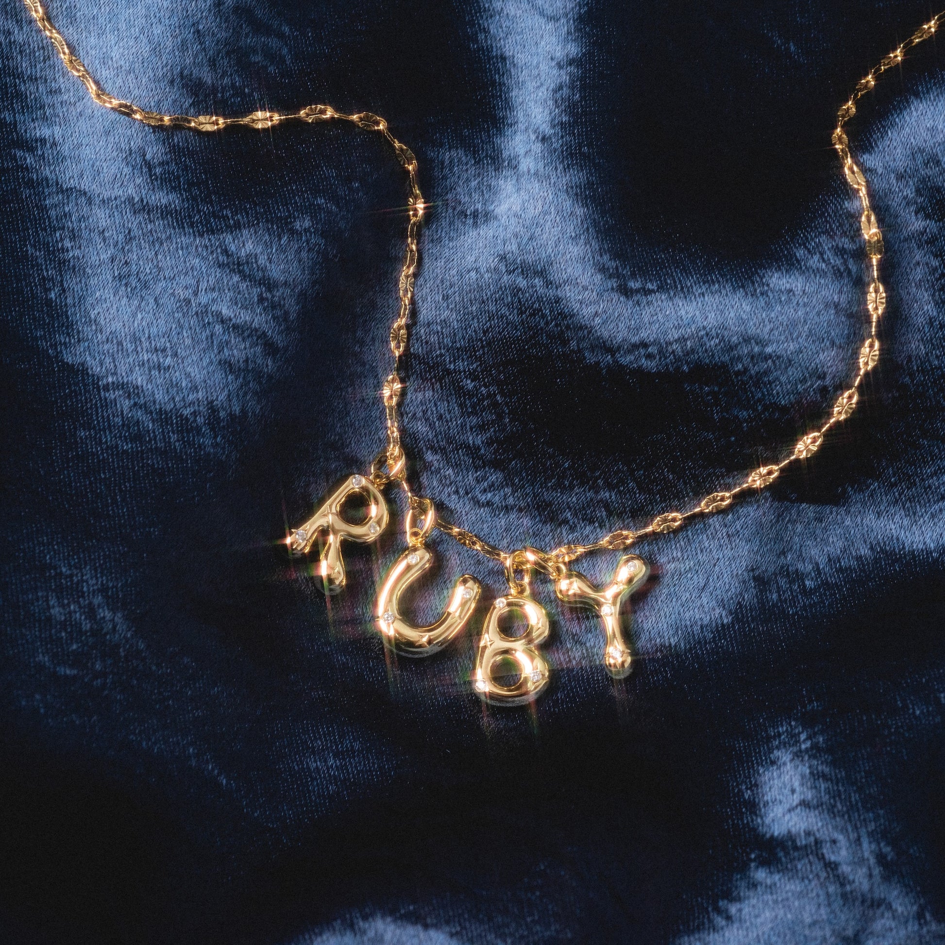 A Star Crossed Celestial Bubble Letter Charm spelling RUBY rests on a dark blue, shiny, textured fabric background.