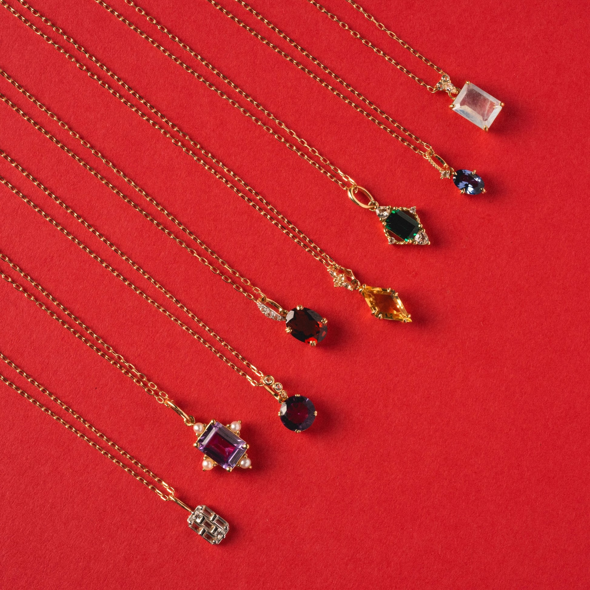 Eight Solid Gold 2025 February Capsule necklaces with unique garnet pendants are arranged diagonally on a bright red background. Each features a distinct shape and color. Set includes a free gift.