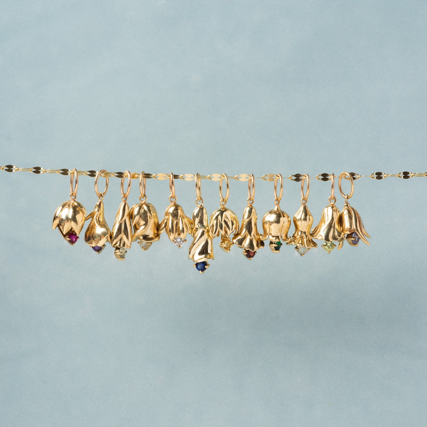 A row of twelve Solid Gold Birth Flower Bud Charms hangs from a gold chain, each shaped like a flower and adorned with colorful gemstones, set against a light blue background.