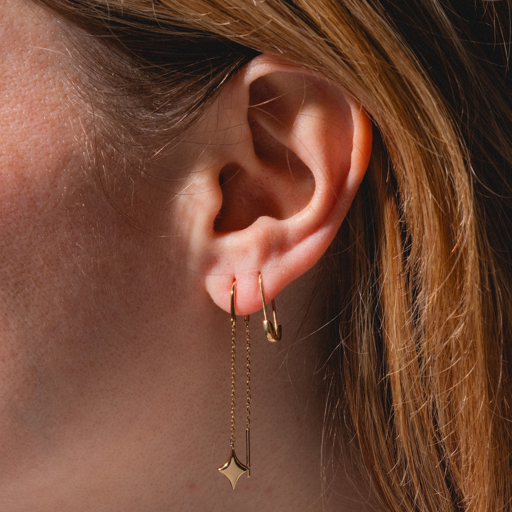 Close-up of a person’s ear with two gold earrings: a small hoop and the Lustra Star Threader Earrings featuring a delicate chain and diamond-shaped charm. Light brown hair frames the ear.
