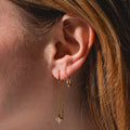 Close-up of a person’s ear with two gold earrings: a small hoop and the Lustra Star Threader Earrings featuring a delicate chain and diamond-shaped charm. Light brown hair frames the ear.