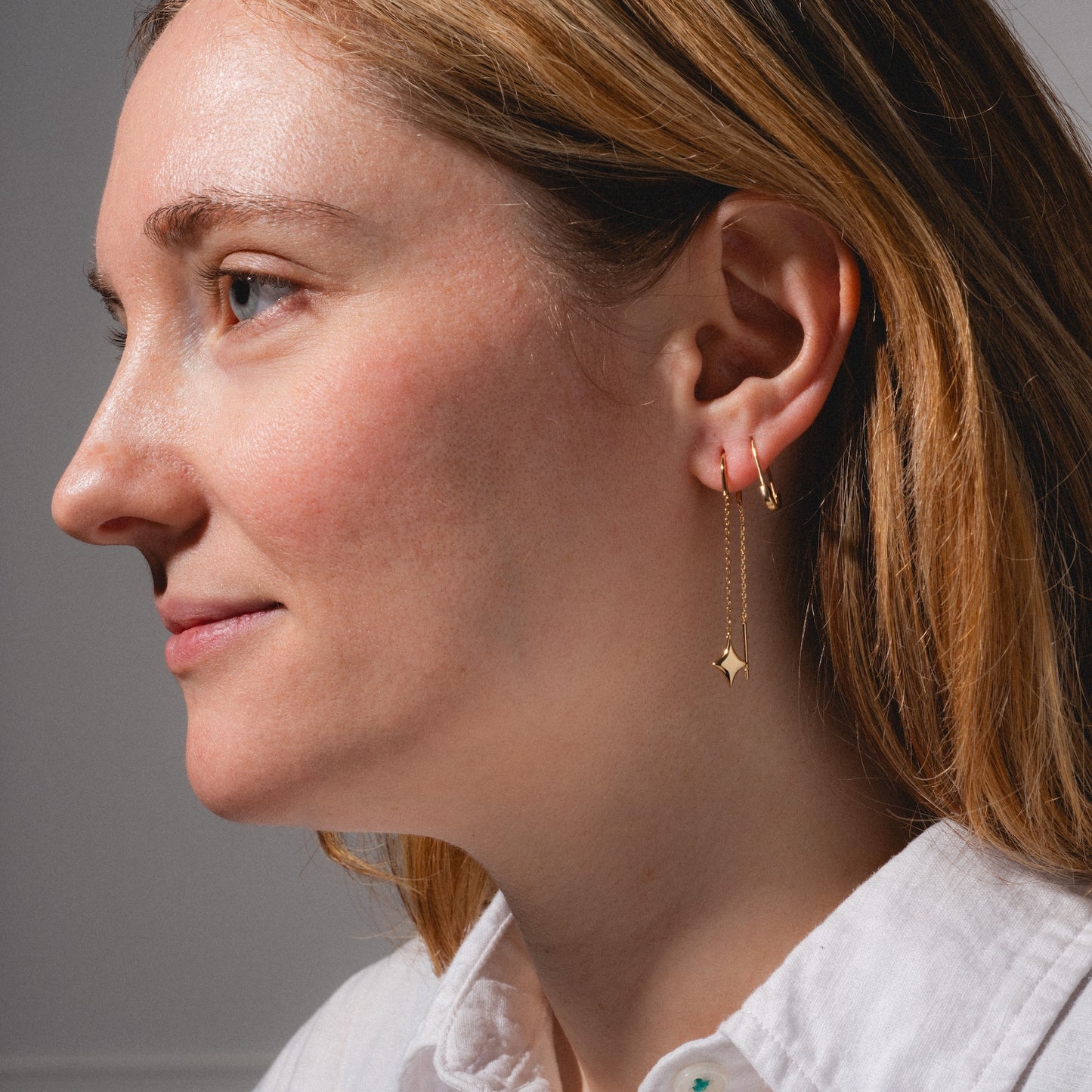 Close-up of a woman with light skin and straight, light brown hair wearing a white collared shirt and Lustra Star Threader Earrings, looking to the left against a neutral background.