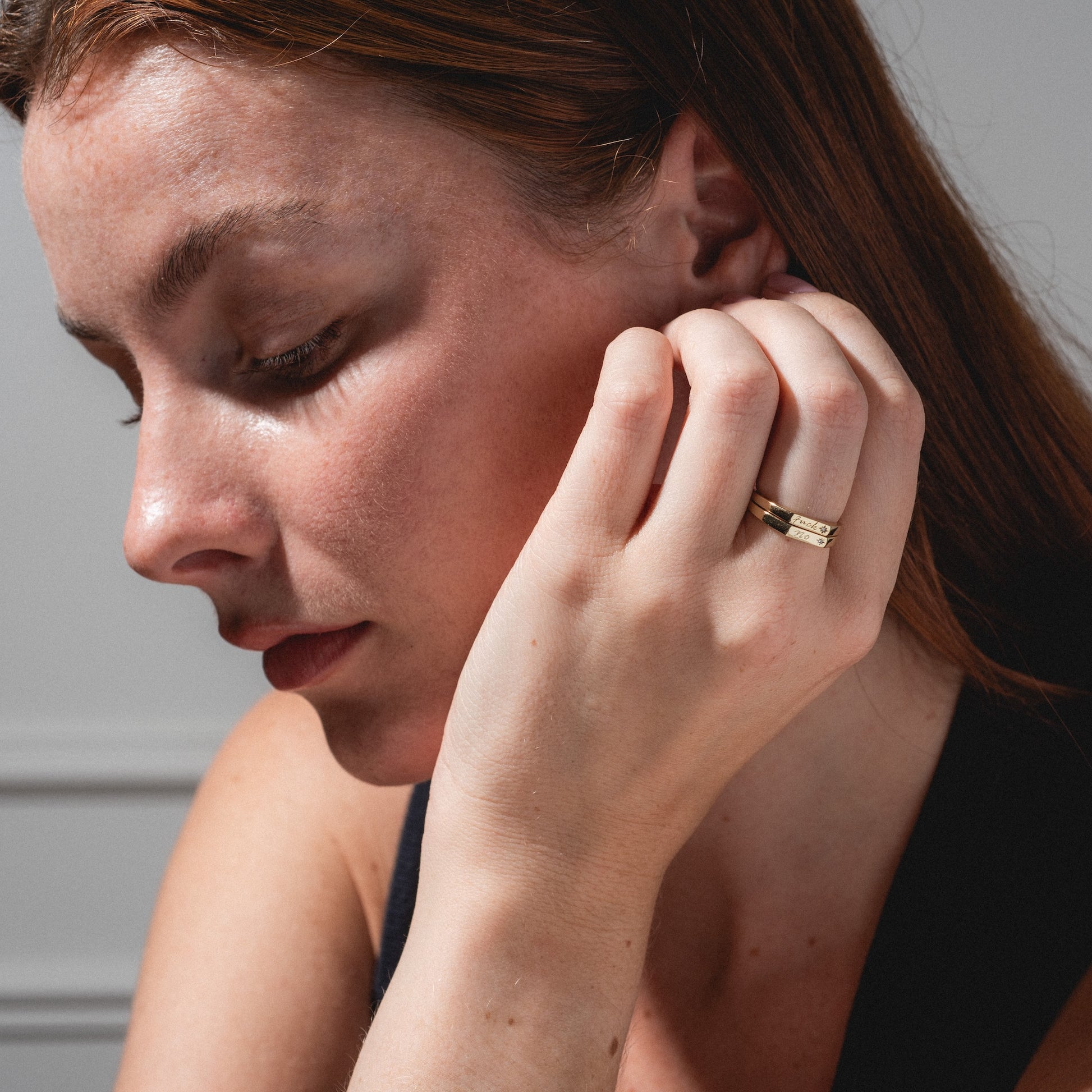 A woman with light skin and brown hair gazes down thoughtfully, her hand near her face displaying two Tiny Conversation Stacking Rings. She wears a black top against a softly lit, neutral background.