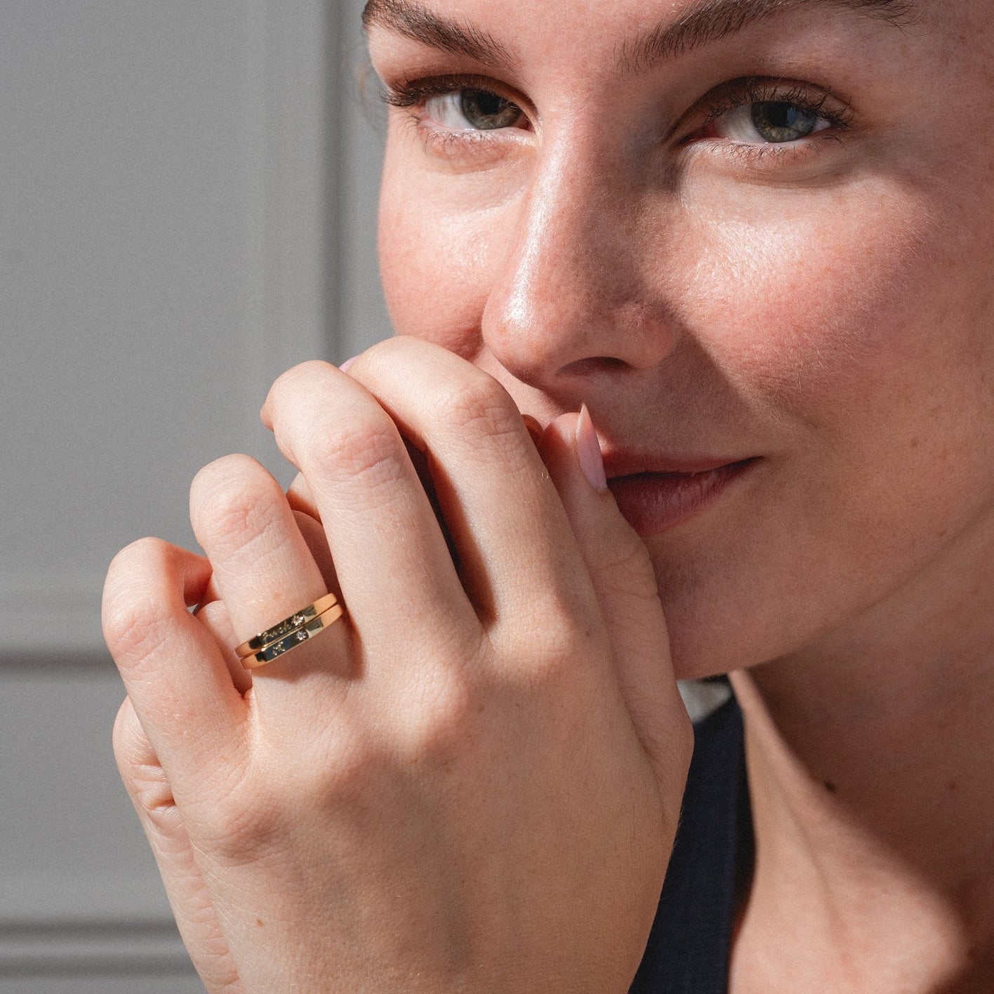 A woman with light skin and brown hair gazes into the camera, her hand covering part of her mouth and showcasing the Tiny Conversation Stacking Ring. She wears a dark sleeveless top, with natural light highlighting her face.