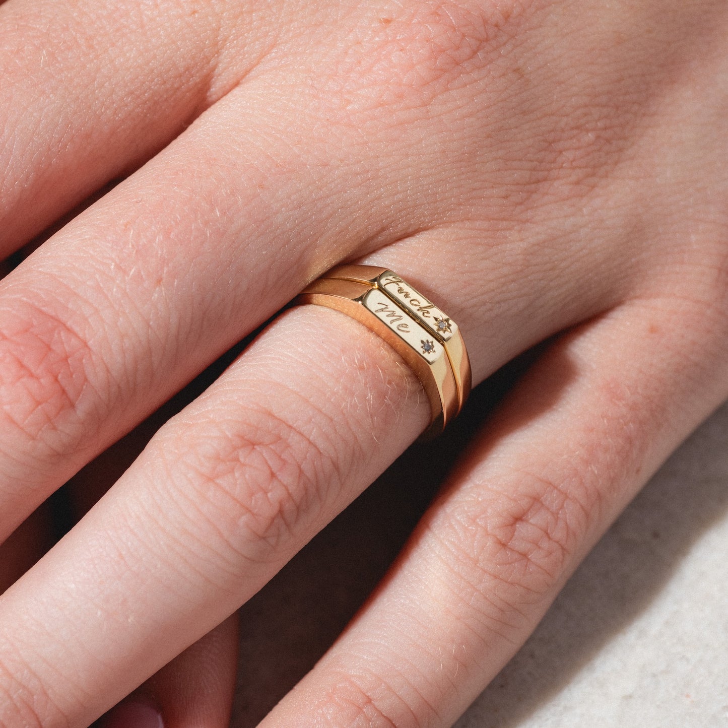 A hand wears three stacked Tiny Conversation Stacking Rings in gold on the middle finger. The top ring is engraved with "made with love" and a small flower. The hand rests on a light surface.