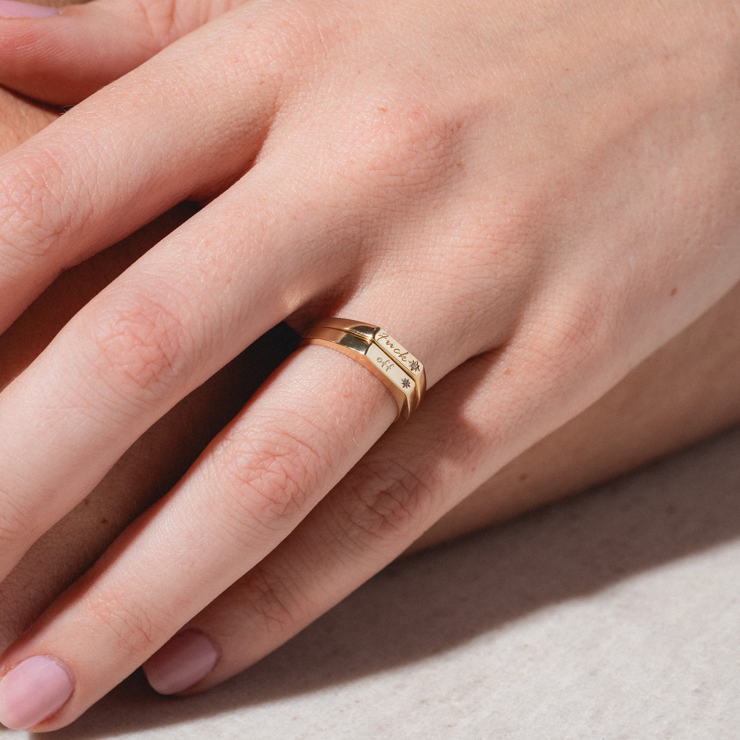 A close-up of a hand wearing two stacked Tiny Conversation Stacking Rings—gold bands with engraved text and star symbols—on the ring finger, set against a neutral background.