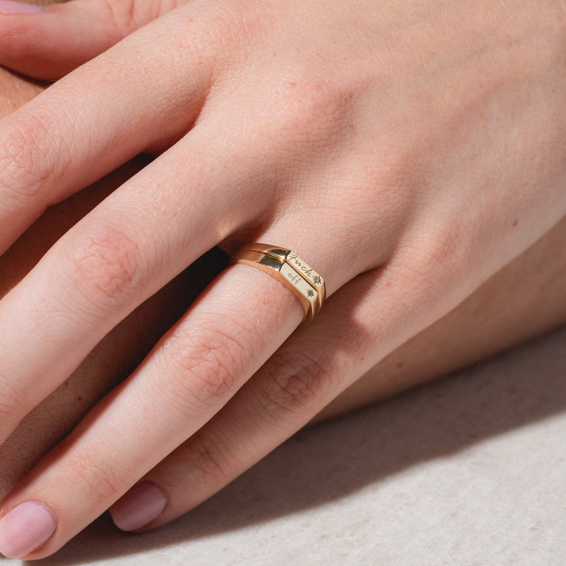 A close-up of a hand wearing two stacked Tiny Conversation Stacking Rings—gold bands with engraved text and star symbols—on the ring finger, set against a neutral background.