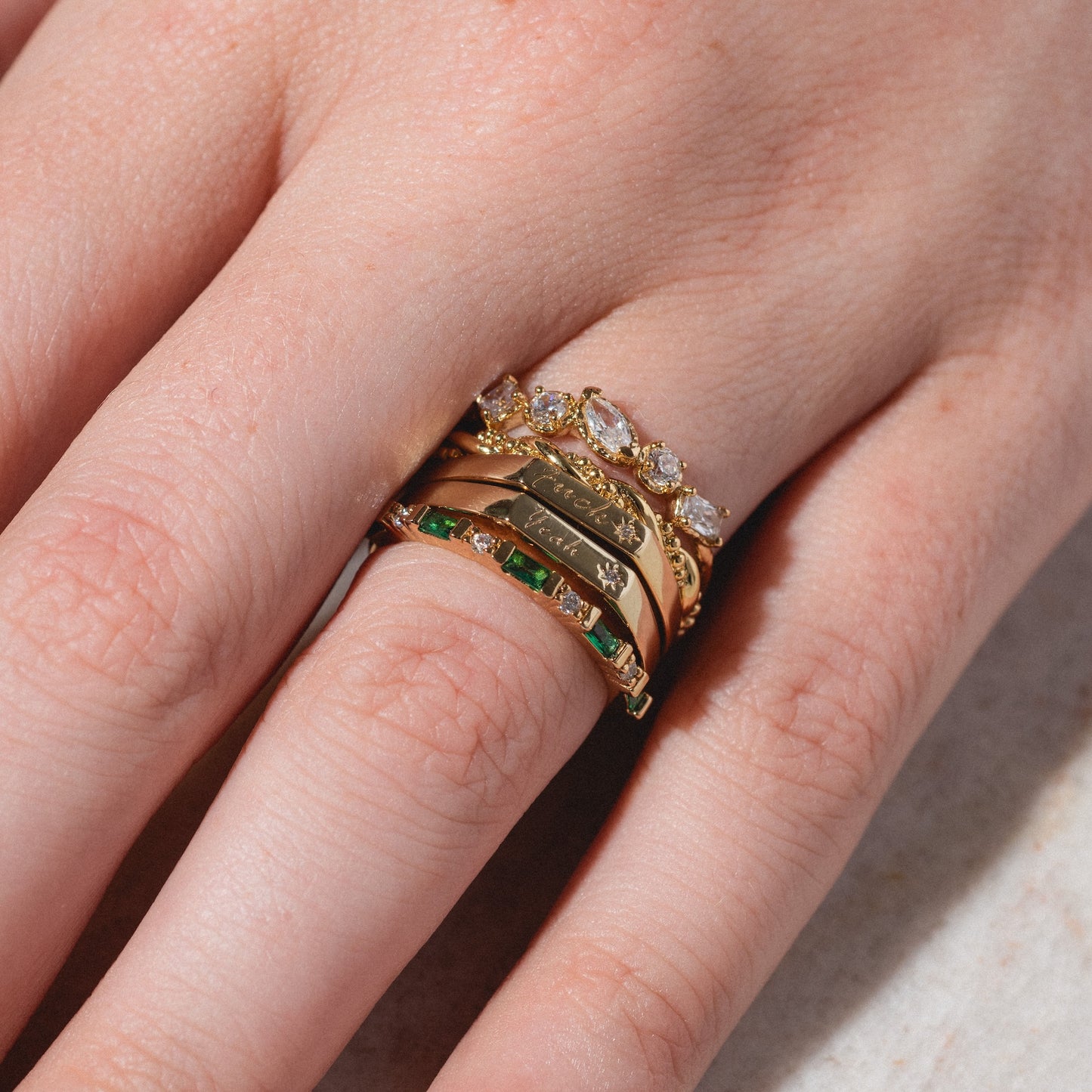A fair-skinned hand wears multiple Tiny Conversation Stacking Rings—gold bands set with small diamonds, pink stones, and green stones—stacked on the ring and middle fingers against a light background.