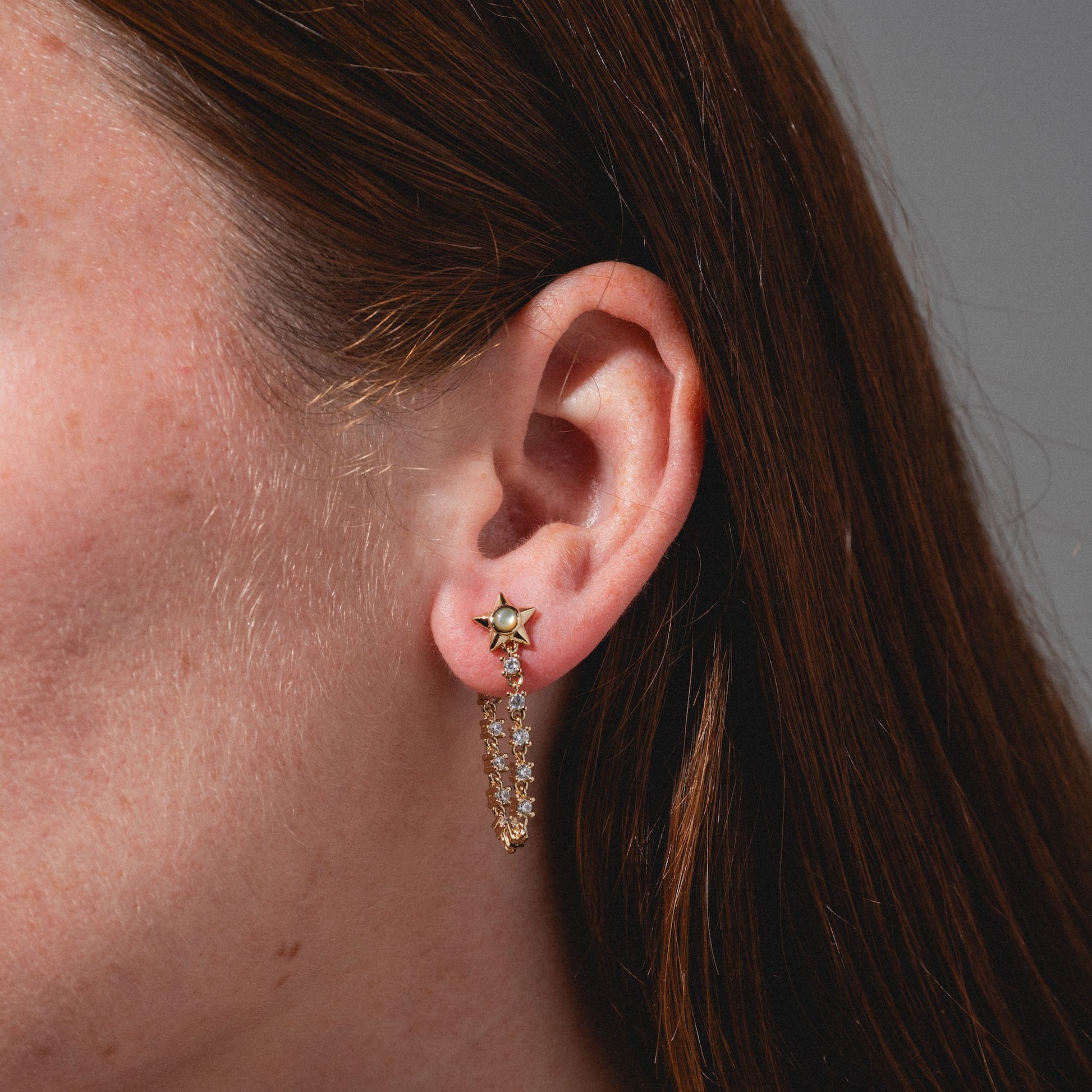 Close-up of an ear with straight brown hair, wearing Lucky Star Tennis Chain Earrings—gold earrings featuring a small star at the lobe and a curved row of sparkling gemstones below.