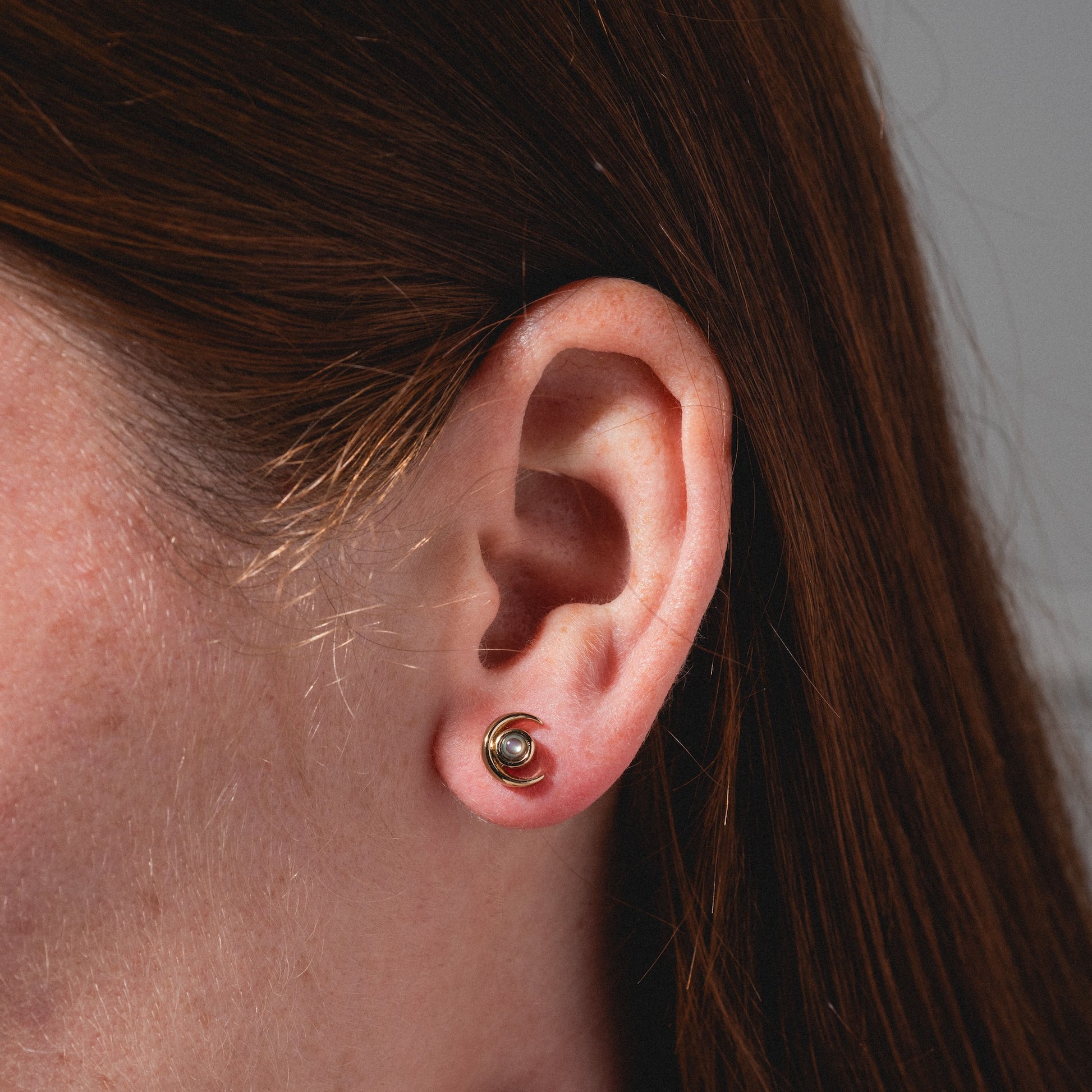 Close-up of a person’s ear with straight brown hair, wearing the Lucky Star and Moon Studs—small, round metallic earrings featuring celestial designs in the earlobe piercing.