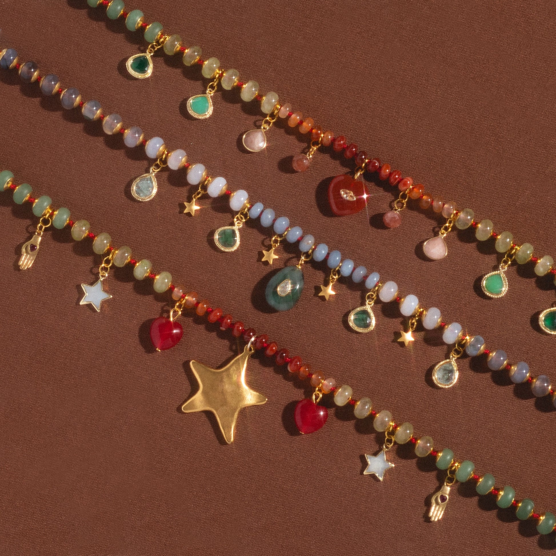 The True North Necklace features colorful beads and various dangling gold charms—stars, hamsa hands, teardrop gems, and heart-shaped pendants—beautifully displayed on a brown surface.