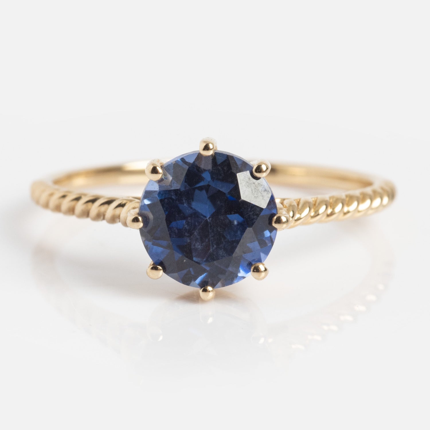 The Solid Gold 2025 December Capsule Classic Tanzanite Statement Ring showcases a round, faceted deep blue tanzanite in a six-prong setting atop a twisted gold band, displayed on a white background.