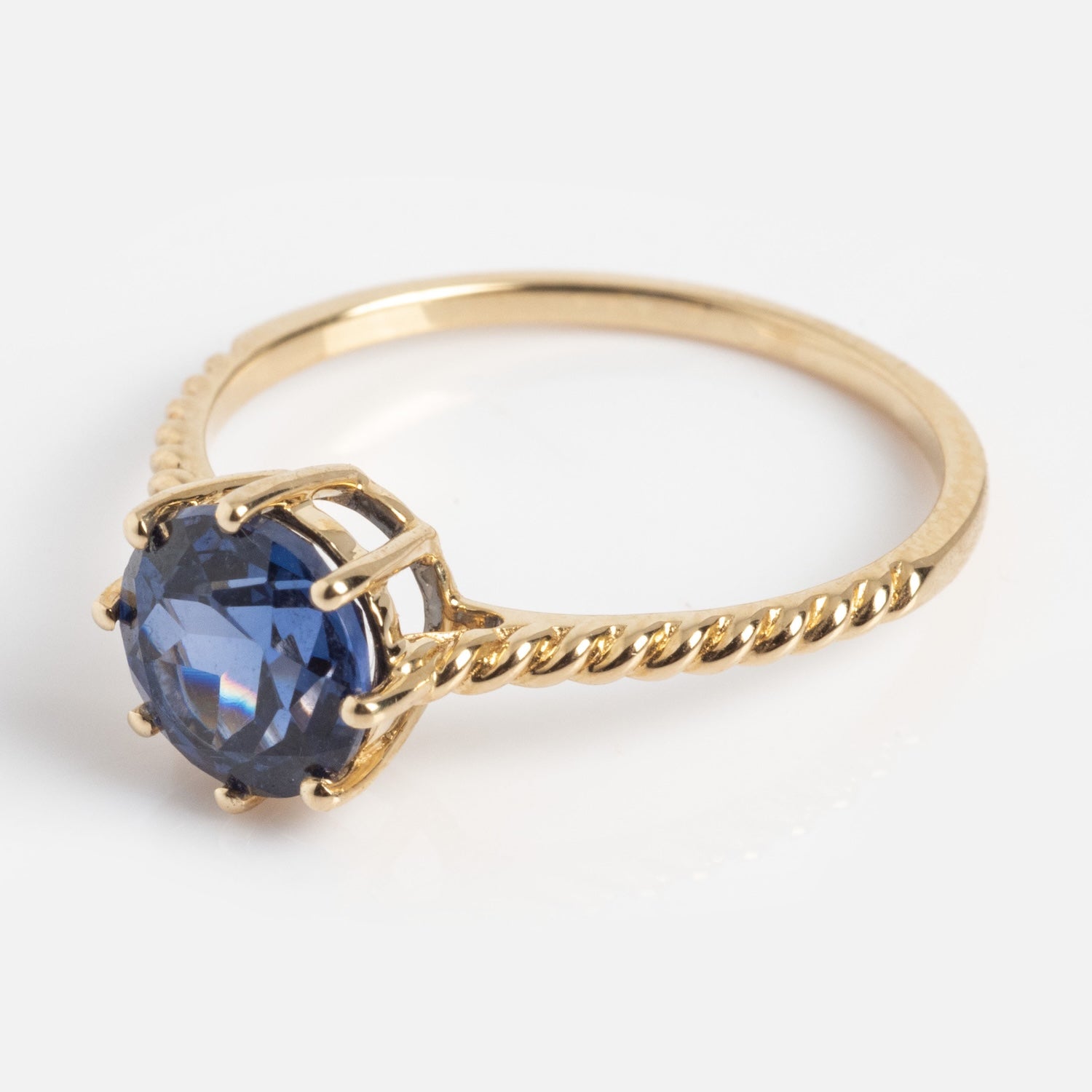 The Solid Gold 2025 December Capsule Classic Tanzanite Statement Ring features a twisted band and a round, dark blue tanzanite gemstone in a prong setting, shown on a white background.