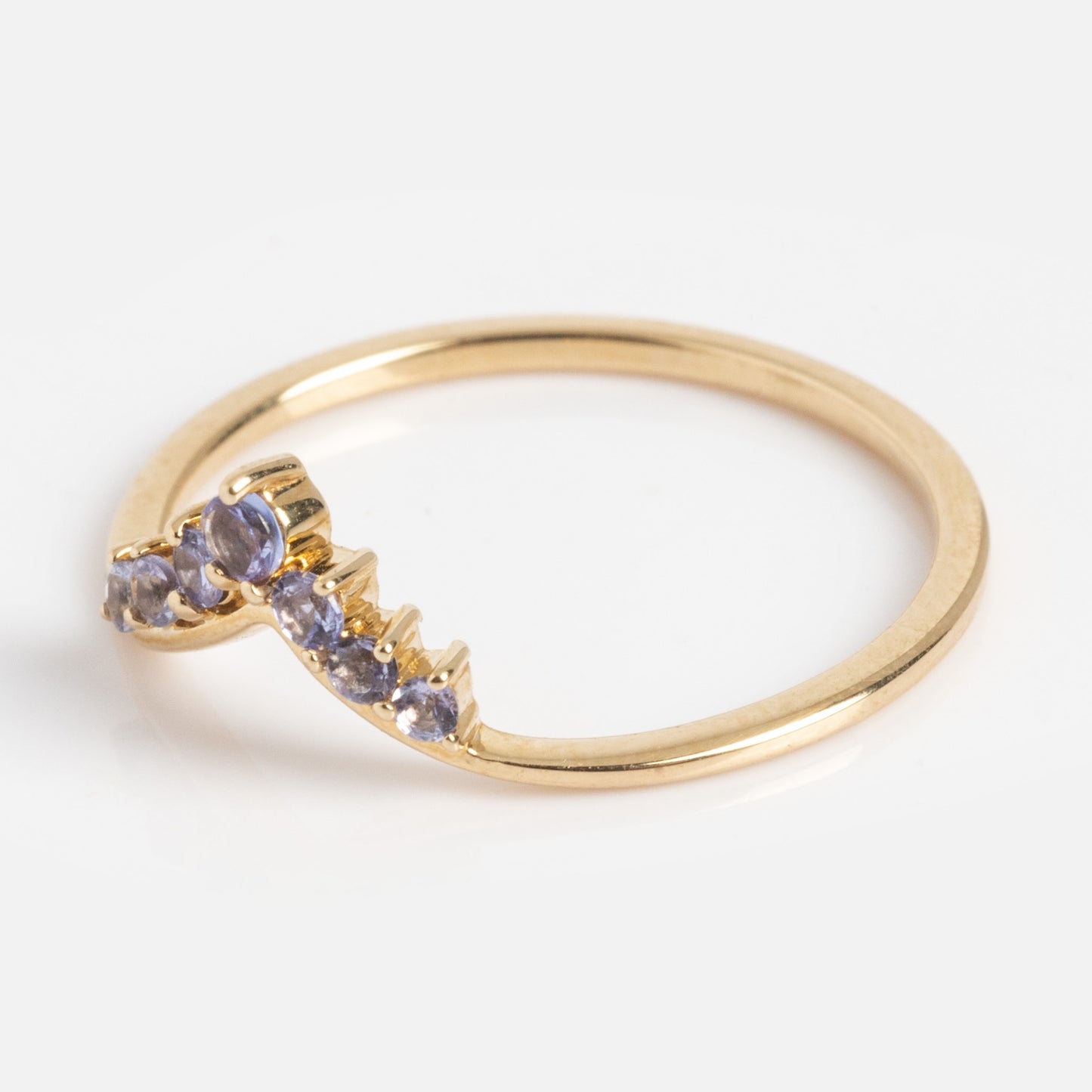 The Solid Gold 2025 December Capsule Delicate Arch Tanzanite Stacking Ring features a slim gold band with a curved row of small, round tanzanite stones, shown on a white background.