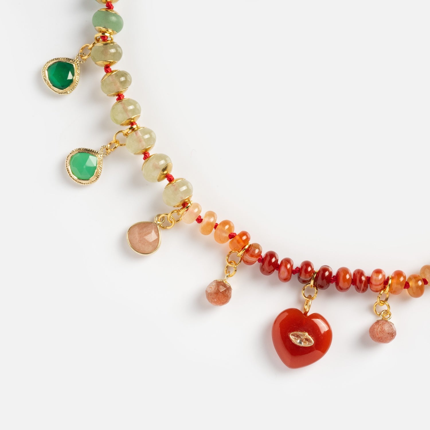 The Golden Hour Necklace features green, yellow, orange, and red beads with gold charms and a central red heart-shaped pendant accented in gold, all displayed on a white background.