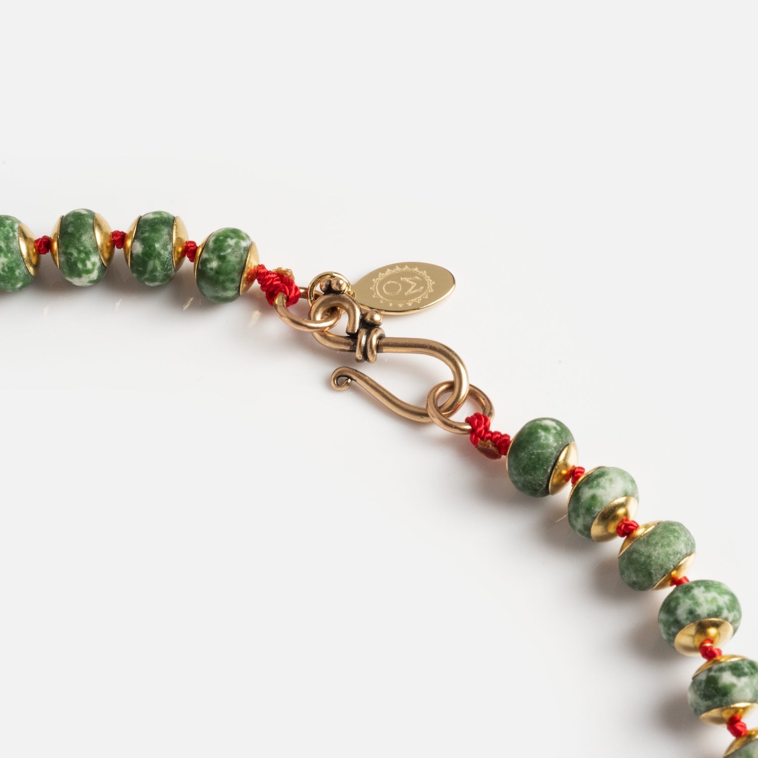 Close-up of the Golden Hour Necklace—a green beaded piece with gold accents, a red cord, gold hook clasp, and a small oval tag engraved with a symbol. Shown against a white background.