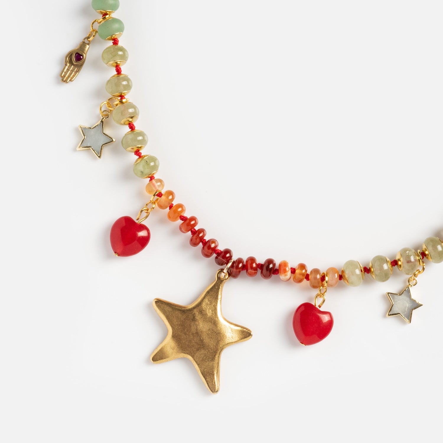 The True North Necklace features red and green beads, gold star and hand charms, silver star charms, and red heart-shaped beads in a playful, eclectic style.