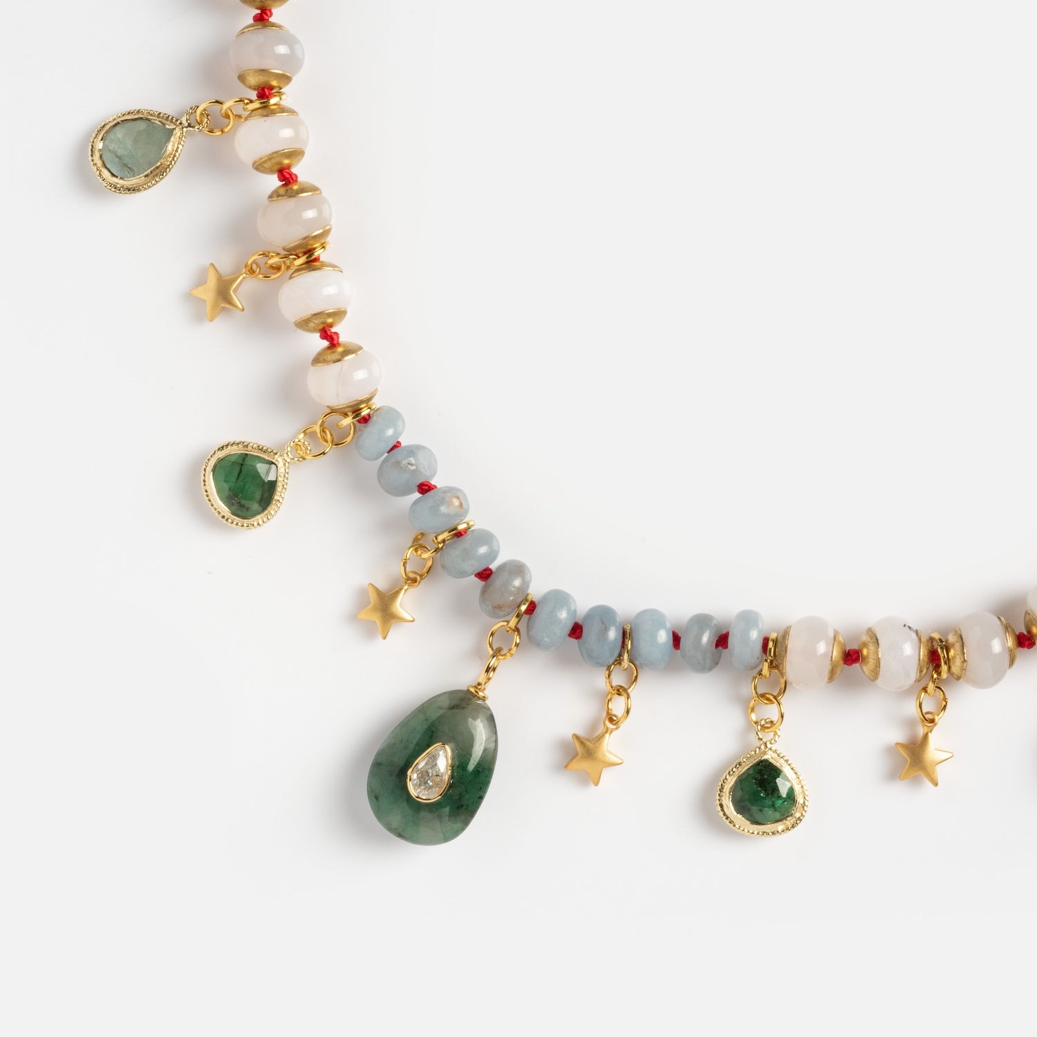 A close-up of the Winter Garden Necklace, featuring green gemstones, gold star charms, gold-bordered teardrop pendants, and white, gray, and red beads against a light background.
