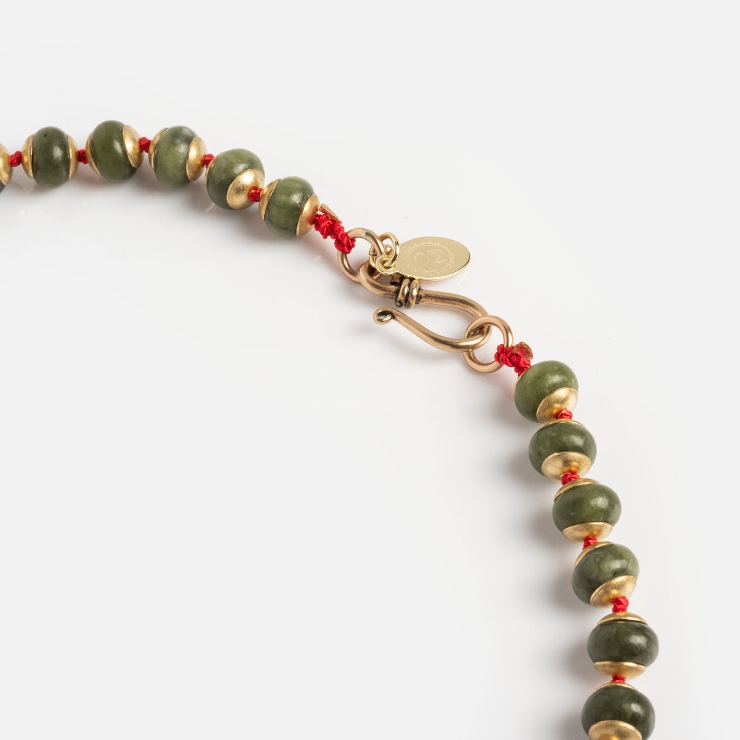 Close-up of the Winter Garden Necklace, featuring green and gold beads on a red cord with a gold hook clasp and a small round gold tag, shown against a white background.