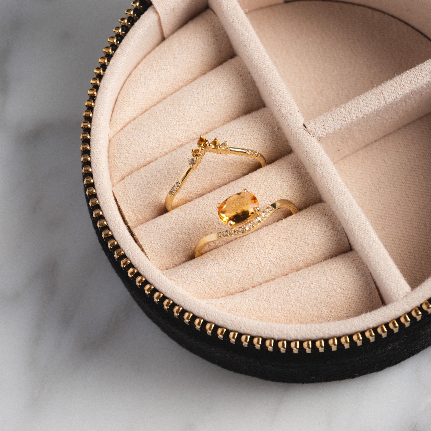 A close-up of the Solid Gold 2025 November Capsule Citrine Ring Set, featuring two gold rings with yellow gemstones in a beige and black zippered jewelry box, displayed on a marble surface; includes a free gift.