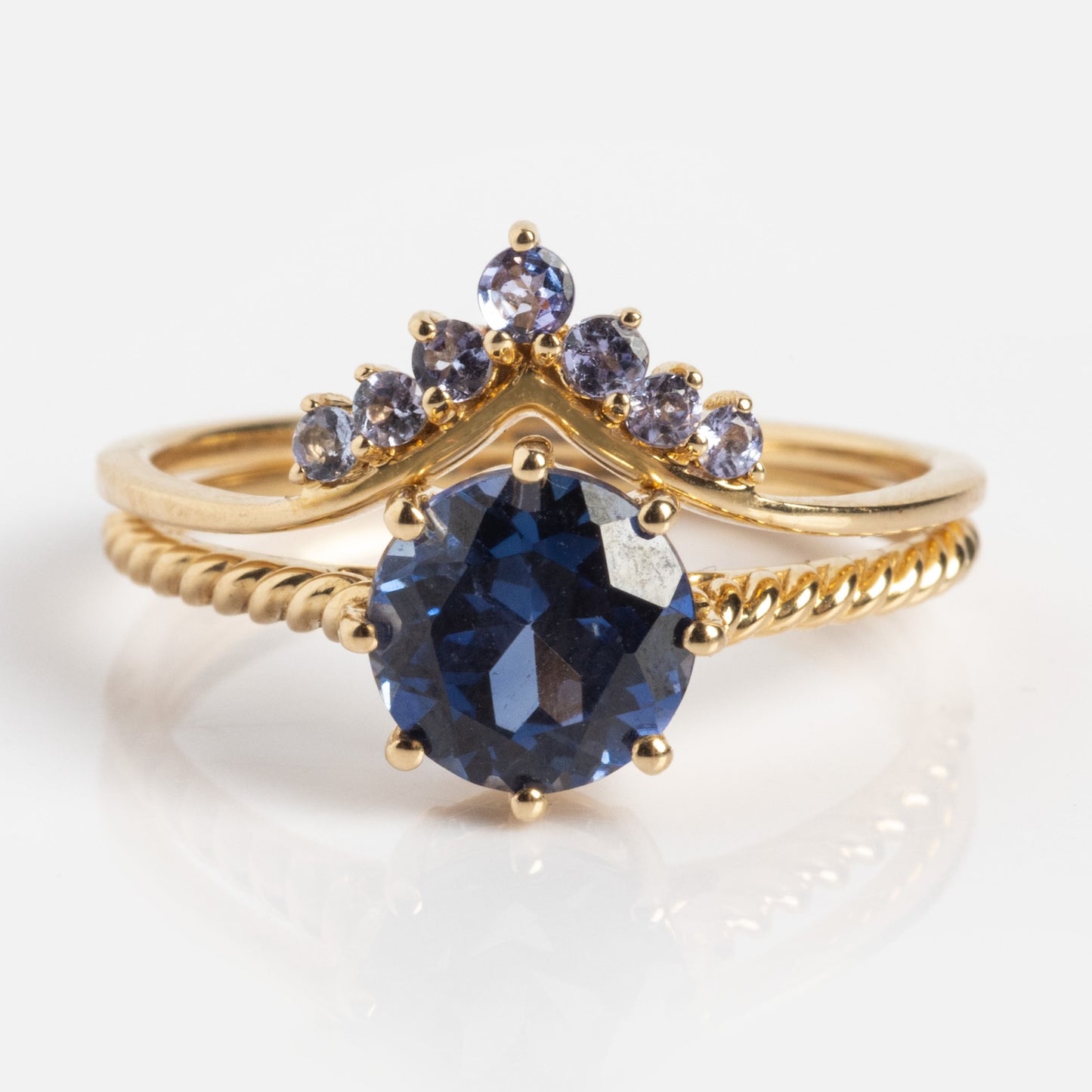 The Solid Gold 2025 December Capsule Tanzanite Ring Set features a large round blue tanzanite ring with a curved band of five small purple stones—both solid gold stacking rings—shown on a white background. Comes with a free gift.