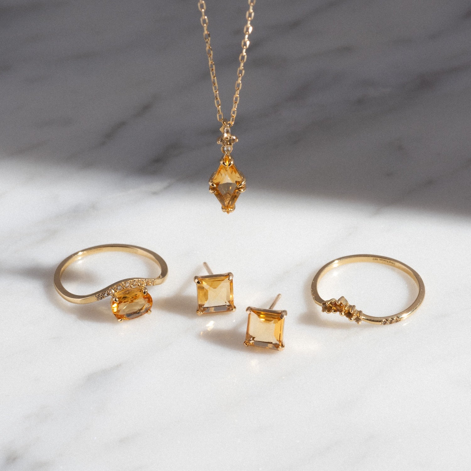 The Solid Gold 2025 November Capsule Art Deco Citrine Pendant Necklace, along with matching rings and square stud earrings set with yellow gemstones, is displayed on a white marble surface with soft lighting.