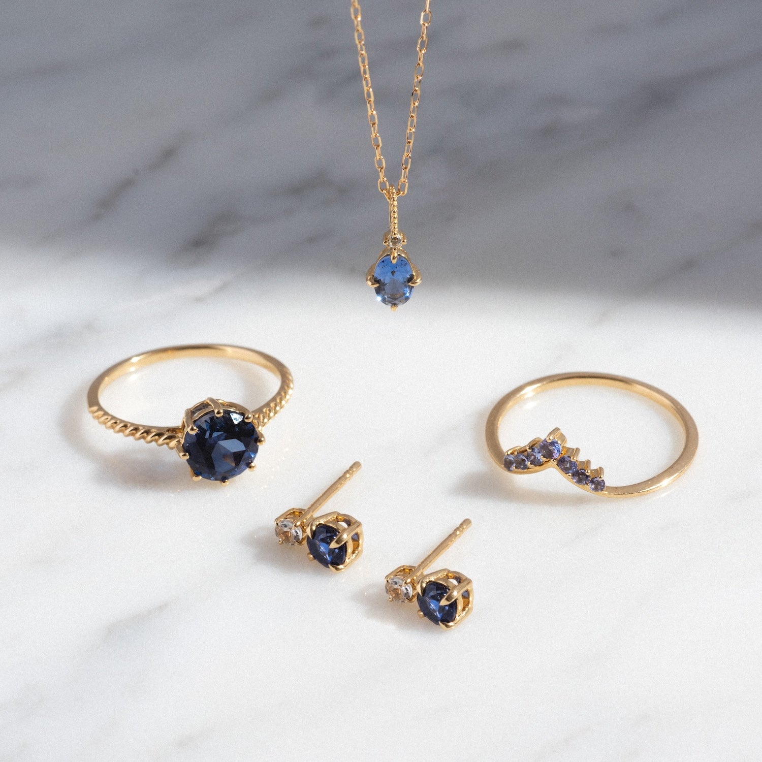 The Solid Gold 2025 December Capsule Classic Tanzanite Pendant Necklace is shown on a marble surface, displayed with coordinating rings and stud earrings in blue gemstone hues, all arranged neatly in natural light.