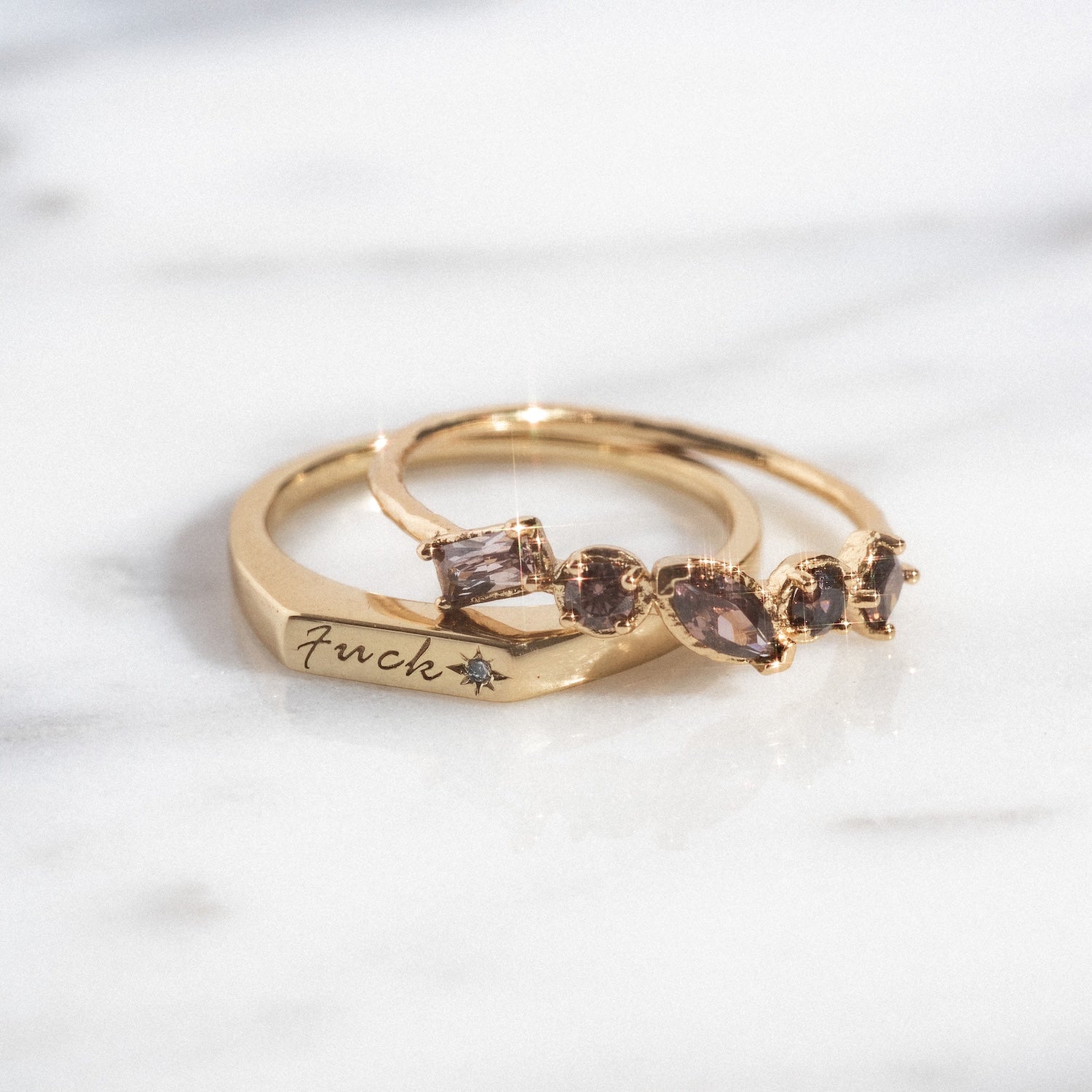 The Tiny Fuck Birthstone Stacking Ring Set features two 14k yellow gold plated rings: one engraved with “F*ck” and a star, the other adorned with four uniquely shaped, light-colored birthstones on marble.