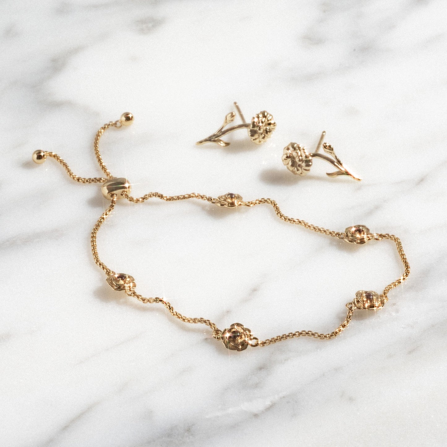The Birth Flower Gift Set, featuring a delicate gold bracelet with round floral accents and a sliding clasp, is displayed on marble beside matching gold floral stud earrings.