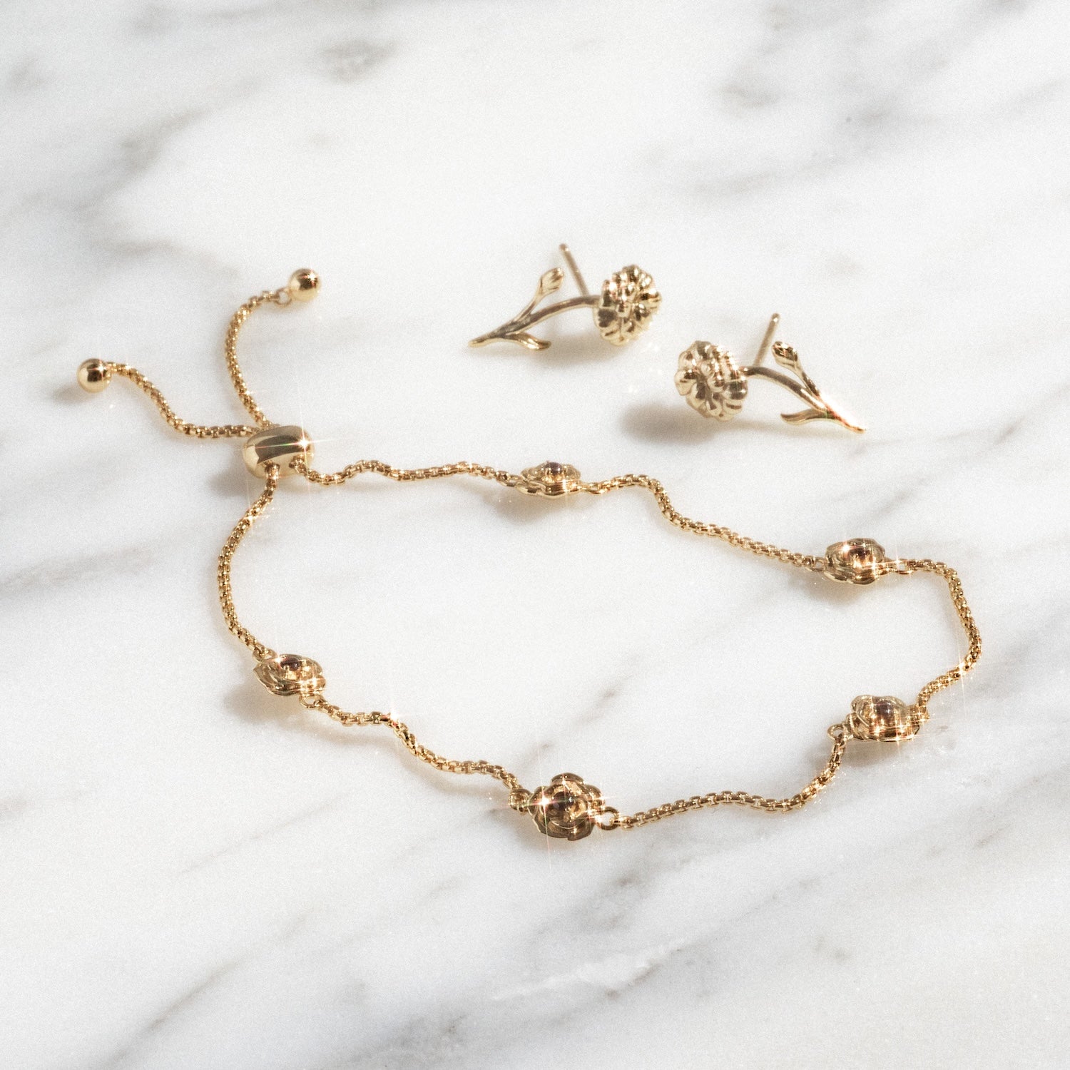 The Birth Flower Gift Set, featuring a delicate gold bracelet with round floral accents and a sliding clasp, is displayed on marble beside matching gold floral stud earrings.