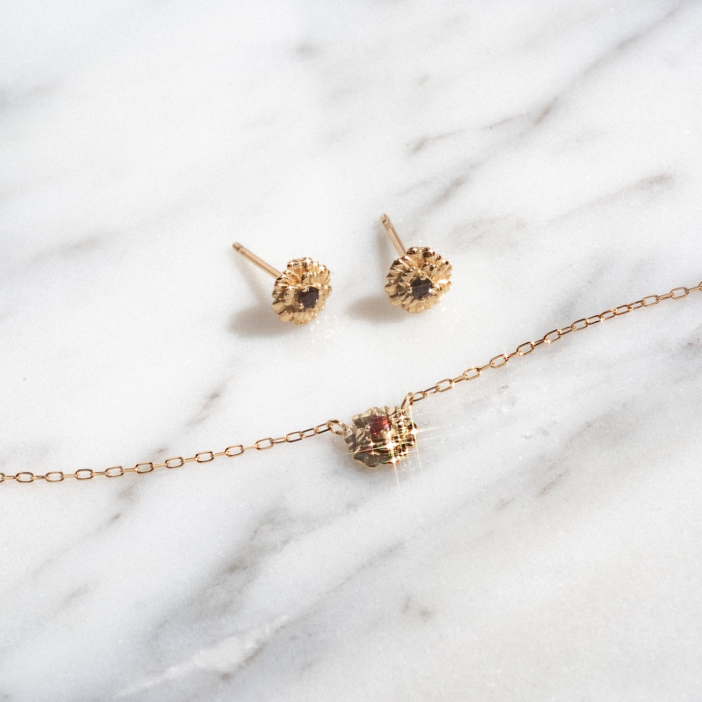 The Solid Gold Birth Flower Gift Set features gold stud earrings with dark central stones and a dainty gold chain necklace with a small red gemstone pendant, all displayed on a white marble surface.